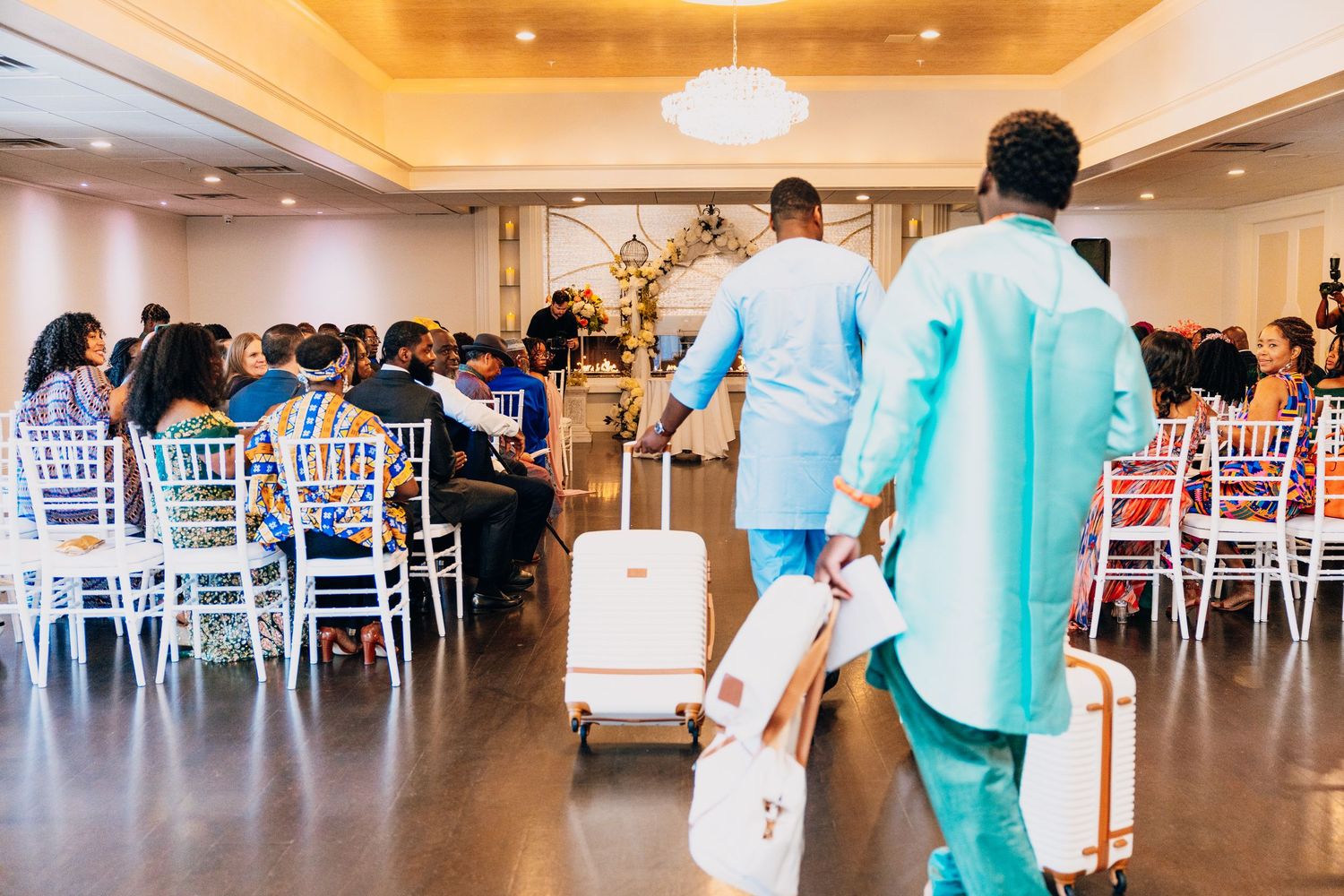Two men in blue traditional attire walk down the aisle at an elegant indoor wedding ceremony with white chairs.