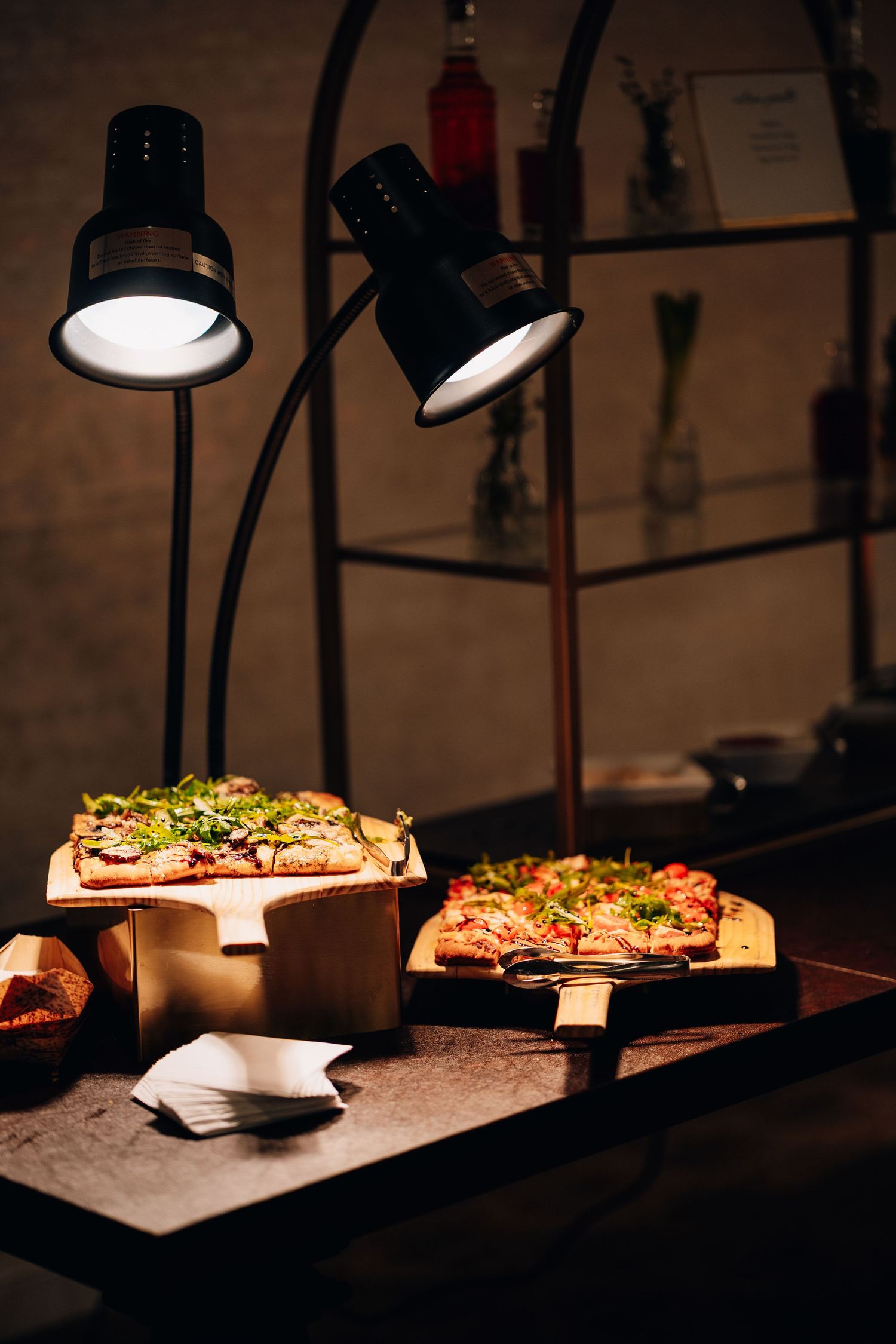 Two flatbread pizzas kept warm under black heat lamps on wooden boards at a dimly lit catering station.
