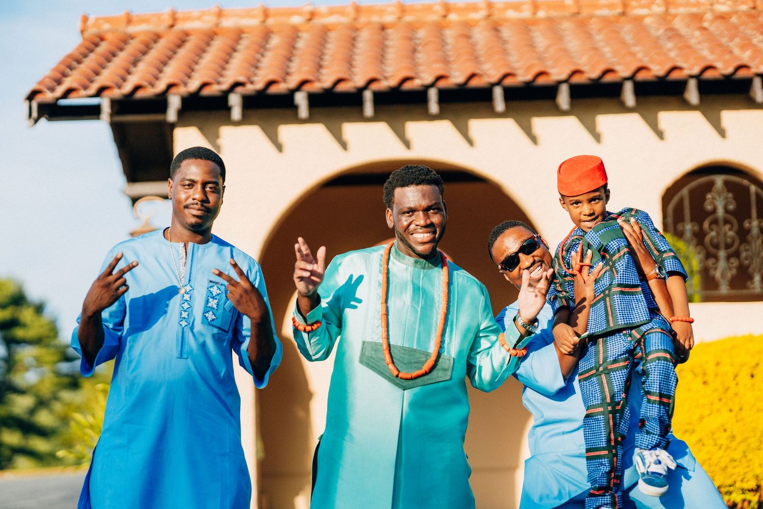 Four men in colorful African traditional attire pose and gesture outdoors.