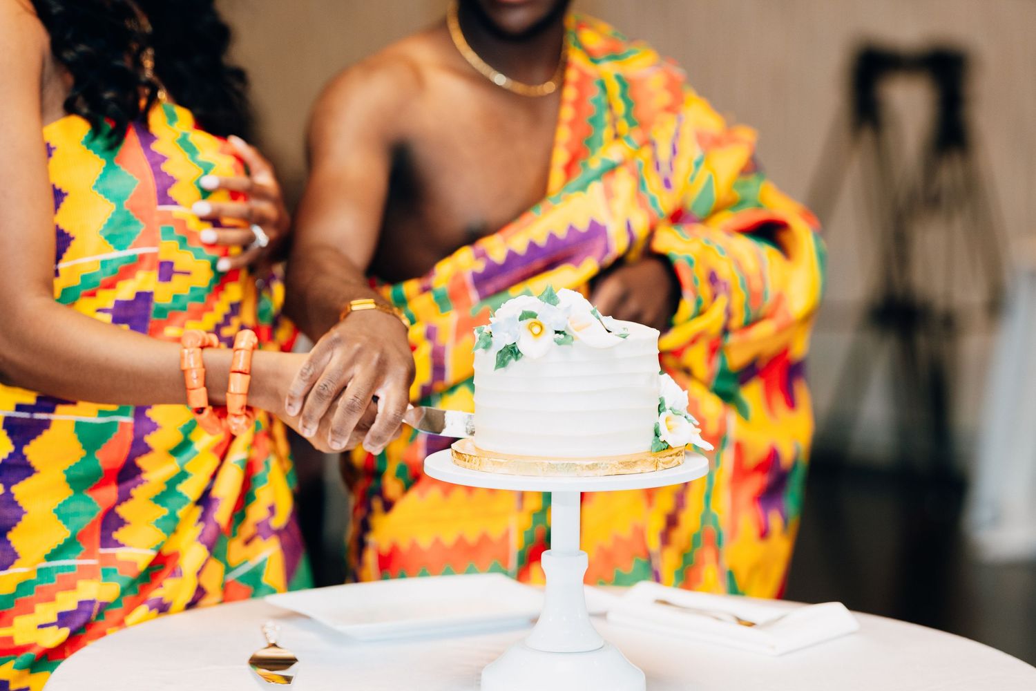 A couple in colorful Kente cloth attire cuts a white wedding cake during a traditional Ghanaian ceremony.