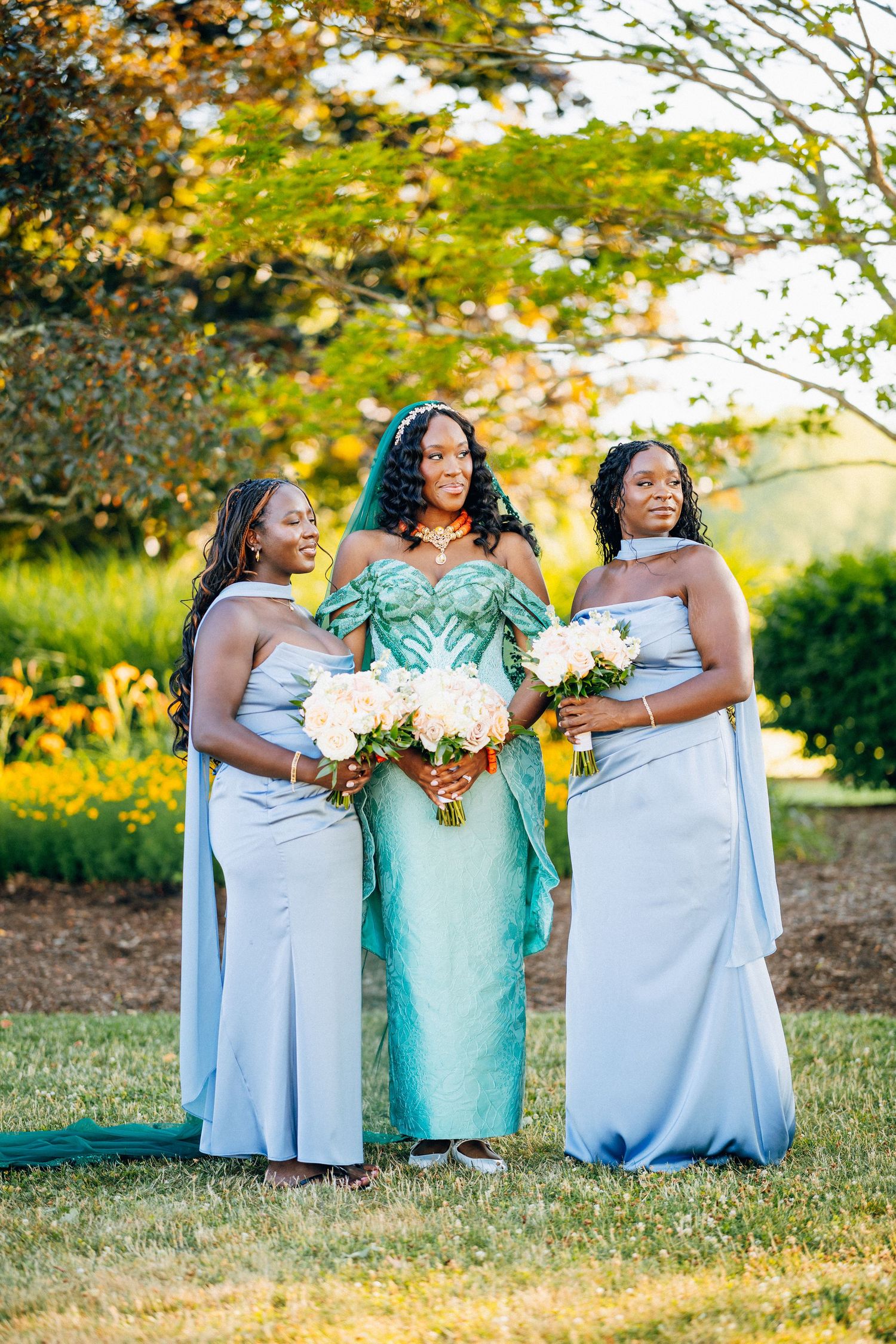 Bride in mint green Nigerian gown stands with two bridesmaids in light blue dresses holding white bouquets.