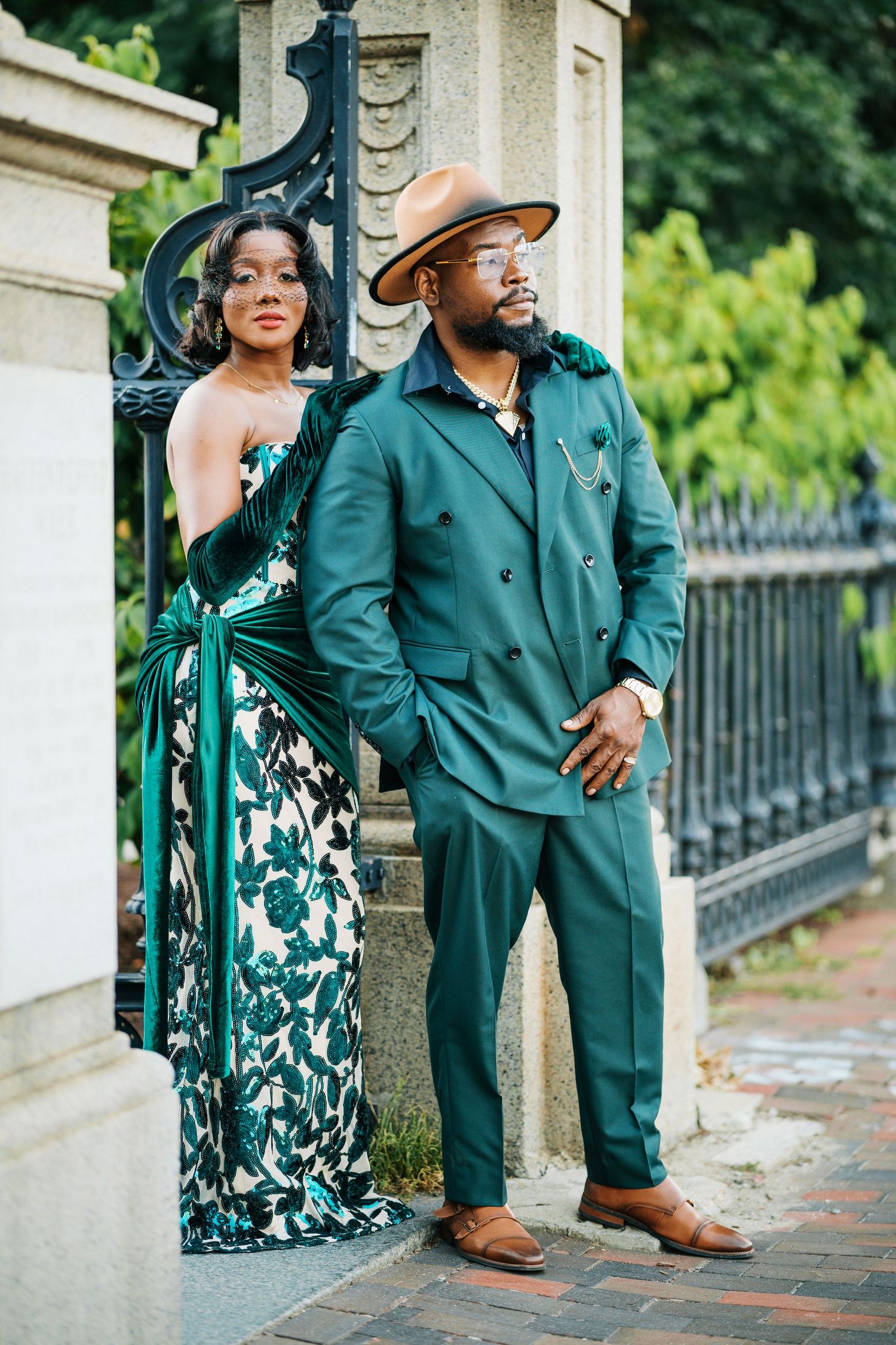 Sophisticated couple posing by ornate iron gates, man in green suit and hat, woman in floral green gown.