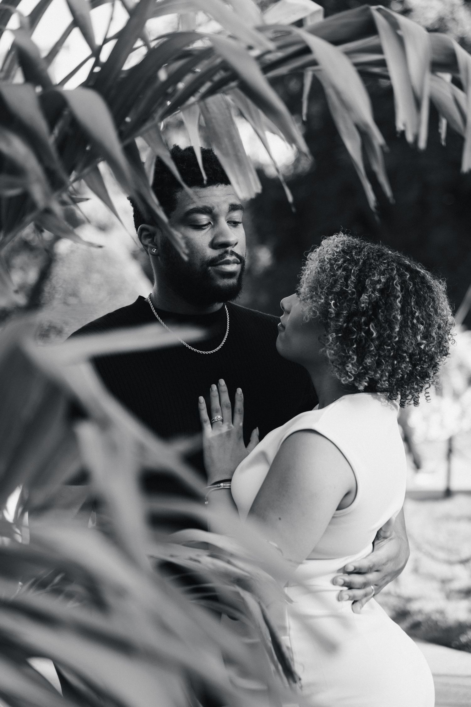 Black and white photo of a couple sharing an intimate moment surrounded by lush tropical foliage.