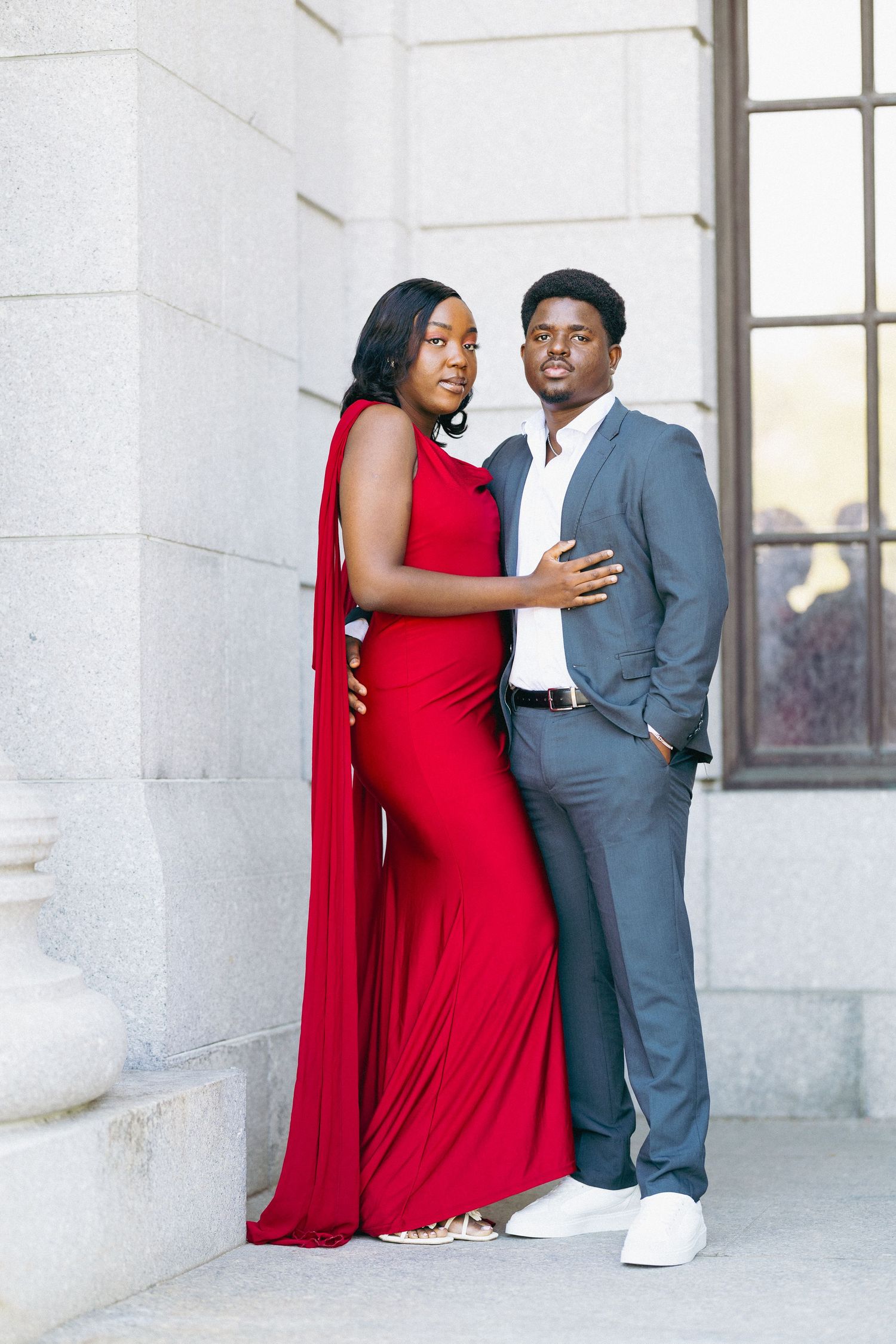 Elegant couple posing outdoors, woman in red cape gown and man in gray suit, against a stone building.