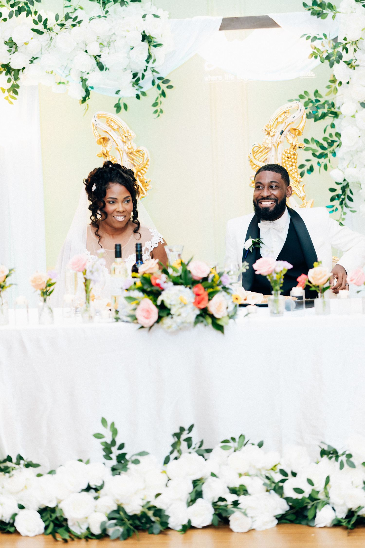Smiling bride and groom seated at a floral-decorated sweetheart table with white roses and greenery at their wedding reception.
