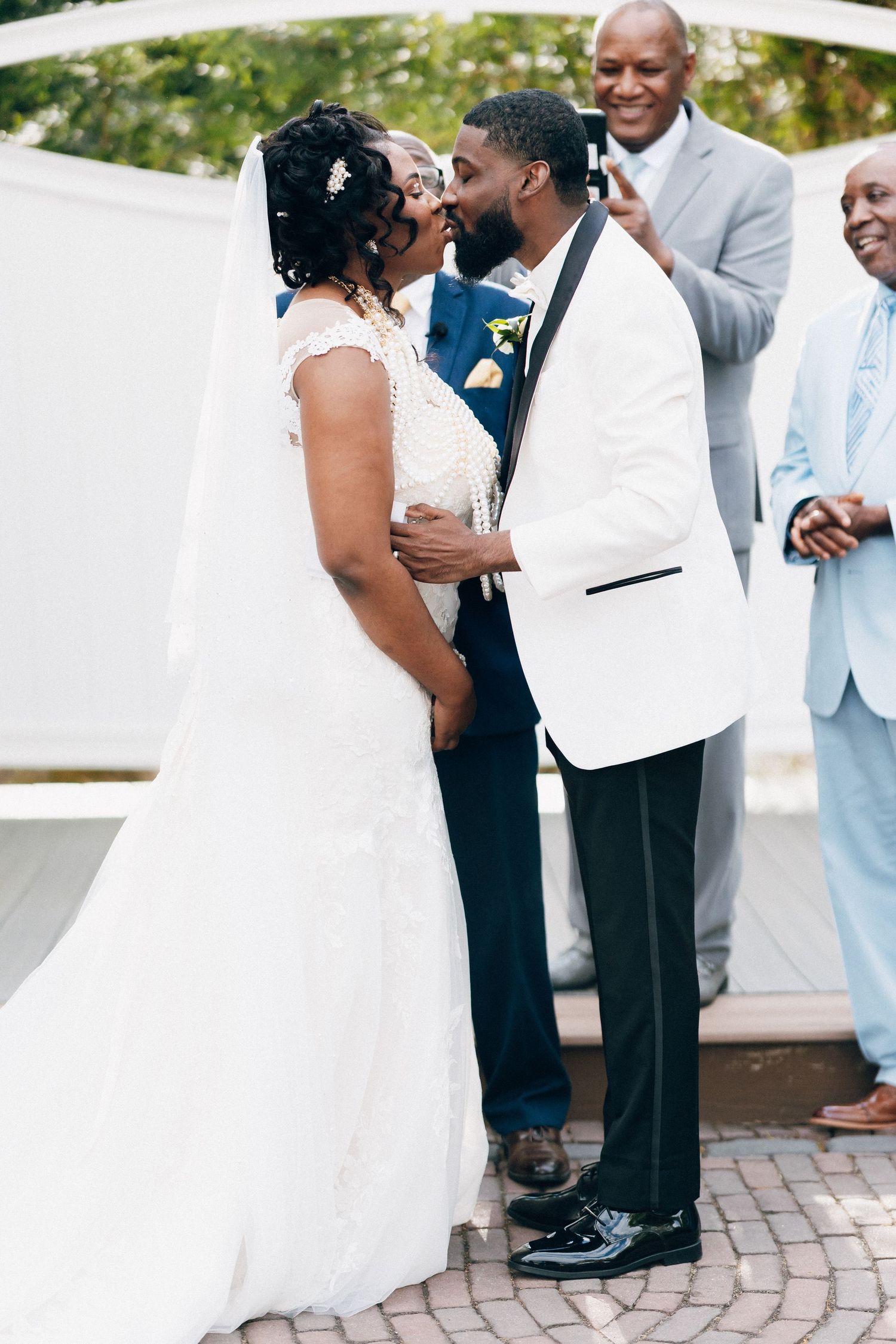 A bride and groom share their first kiss at an outdoor wedding ceremony, surrounded by smiling guests.