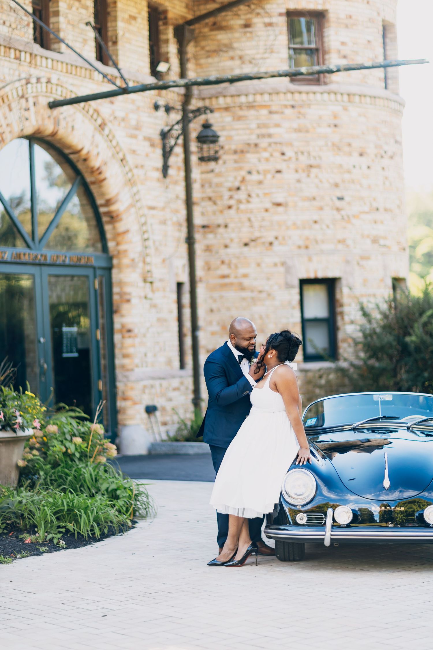 Bride and groom sharing a romantic moment beside a classic black car at a brick venue.