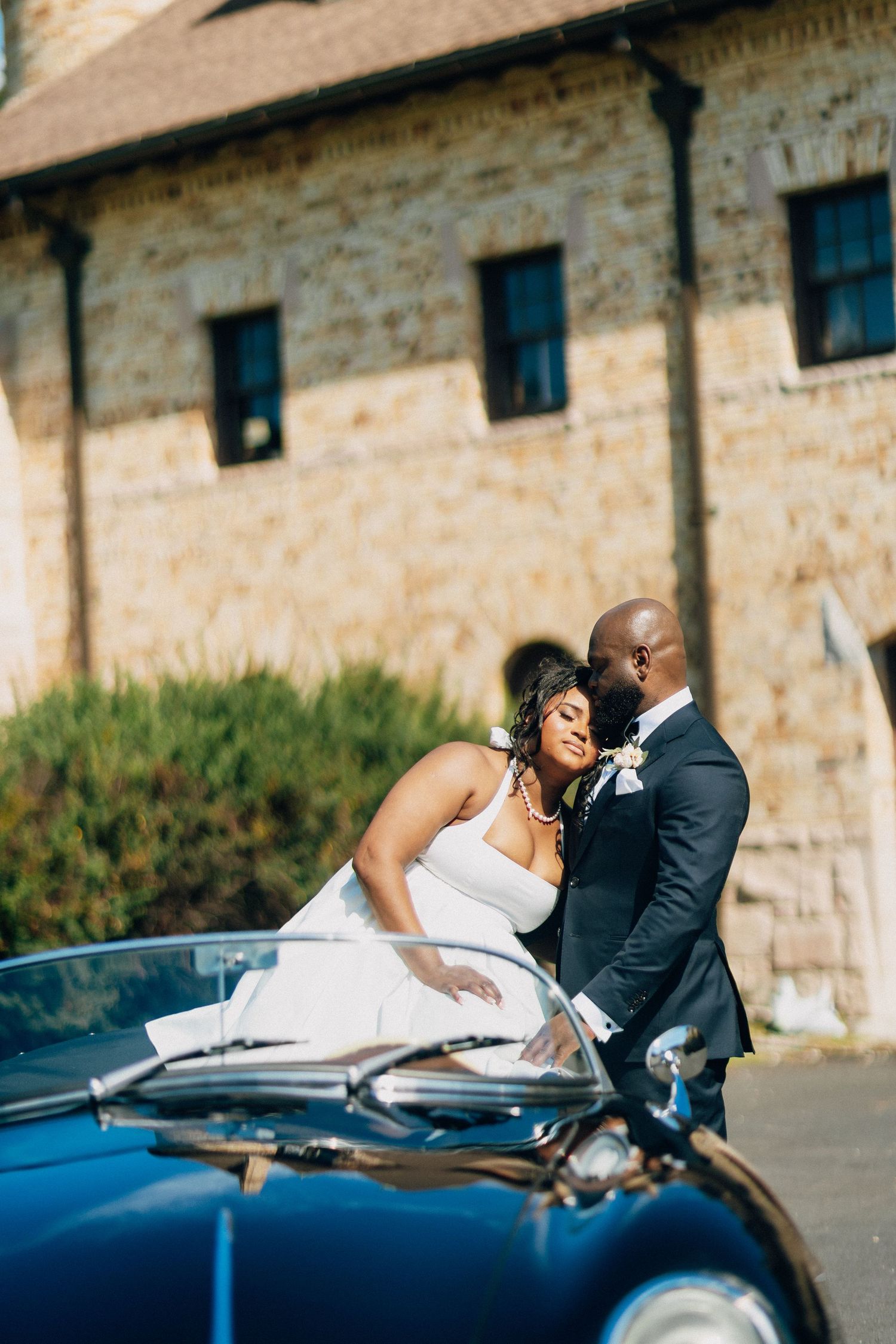African American bride and groom embracing beside a vintage blue Porsche at their wedding.