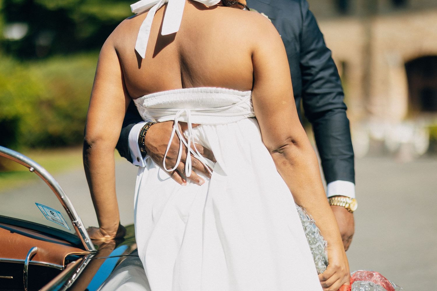 A bride in a halter-back white dress holds hands with her groom beside a vintage car.