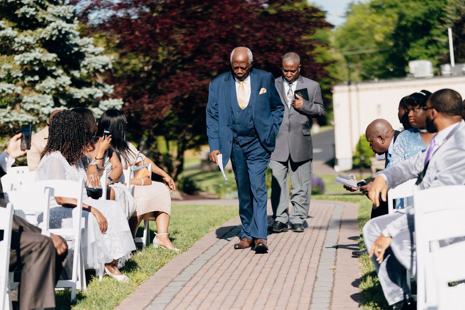 A man in a blue suit walks down an outdoor wedding aisle lined with white chairs and guests on a sunny day.