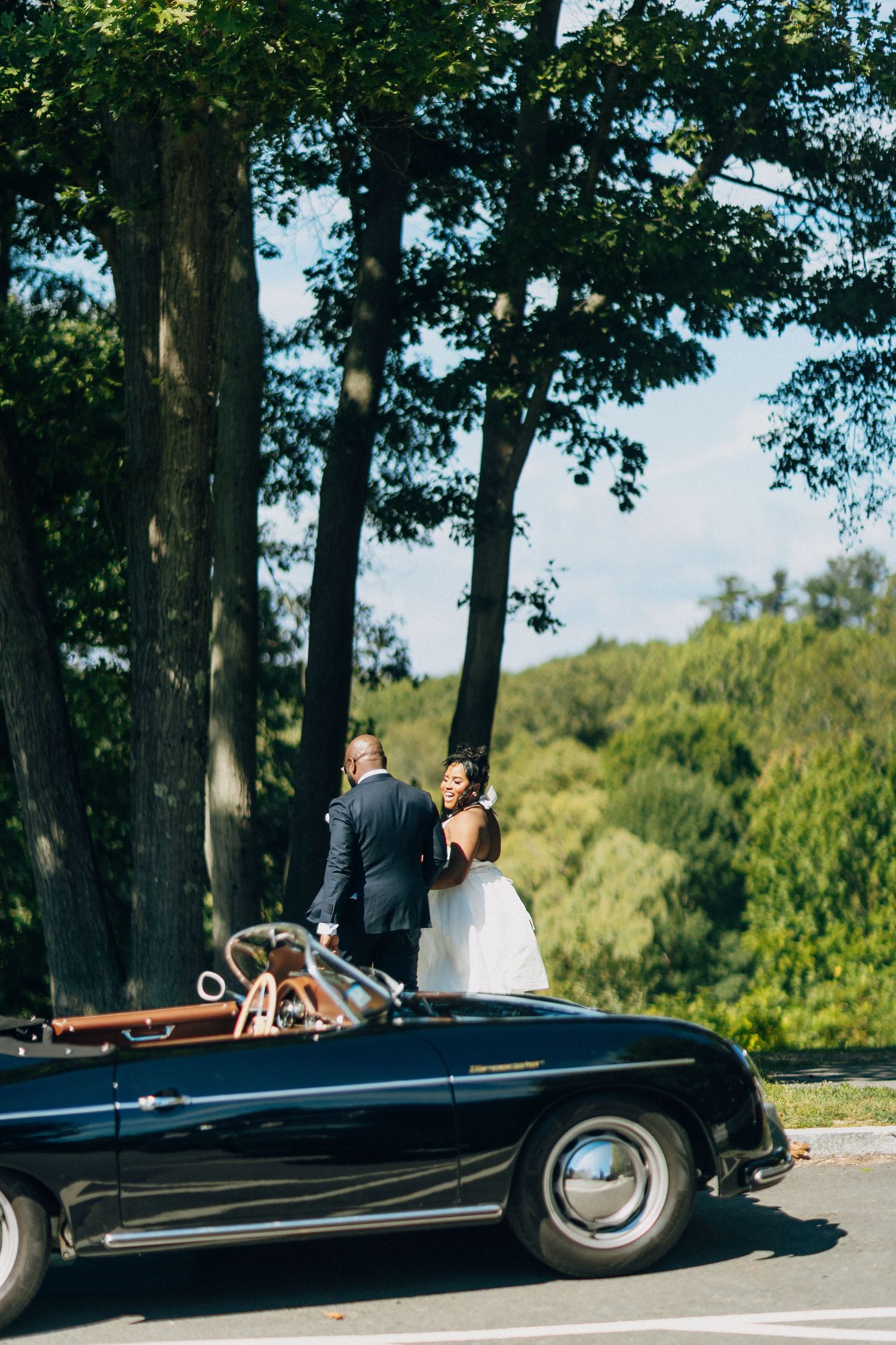 Bride and groom standing together beside a classic black vintage convertible under tall trees.