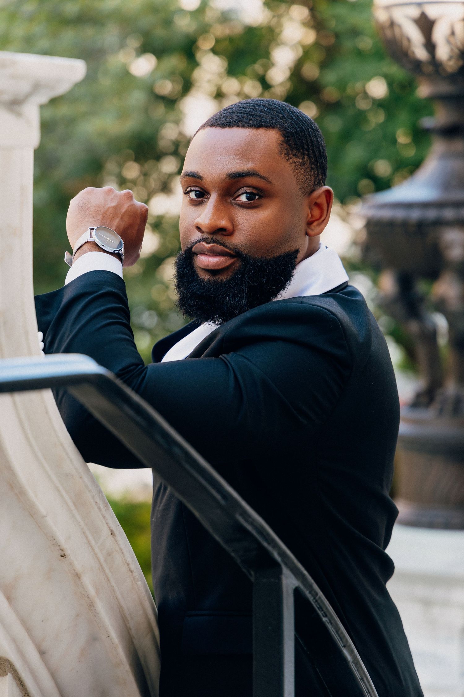 Stylish Black man in a black suit with a beard and watch posing confidently outdoors near a stone fountain.