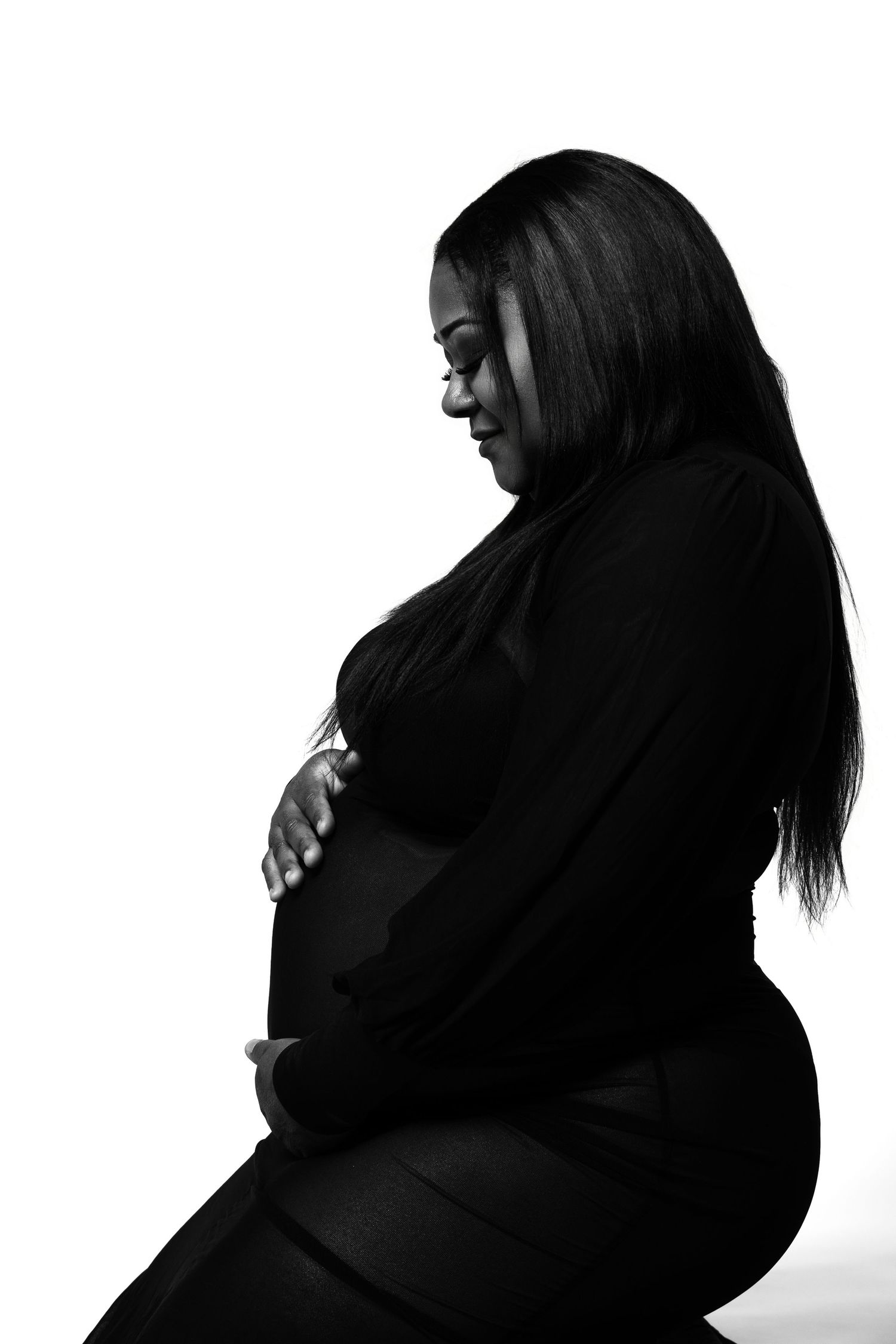 Black and white profile portrait of a pregnant woman in black clothing cradling her baby bump against white background.