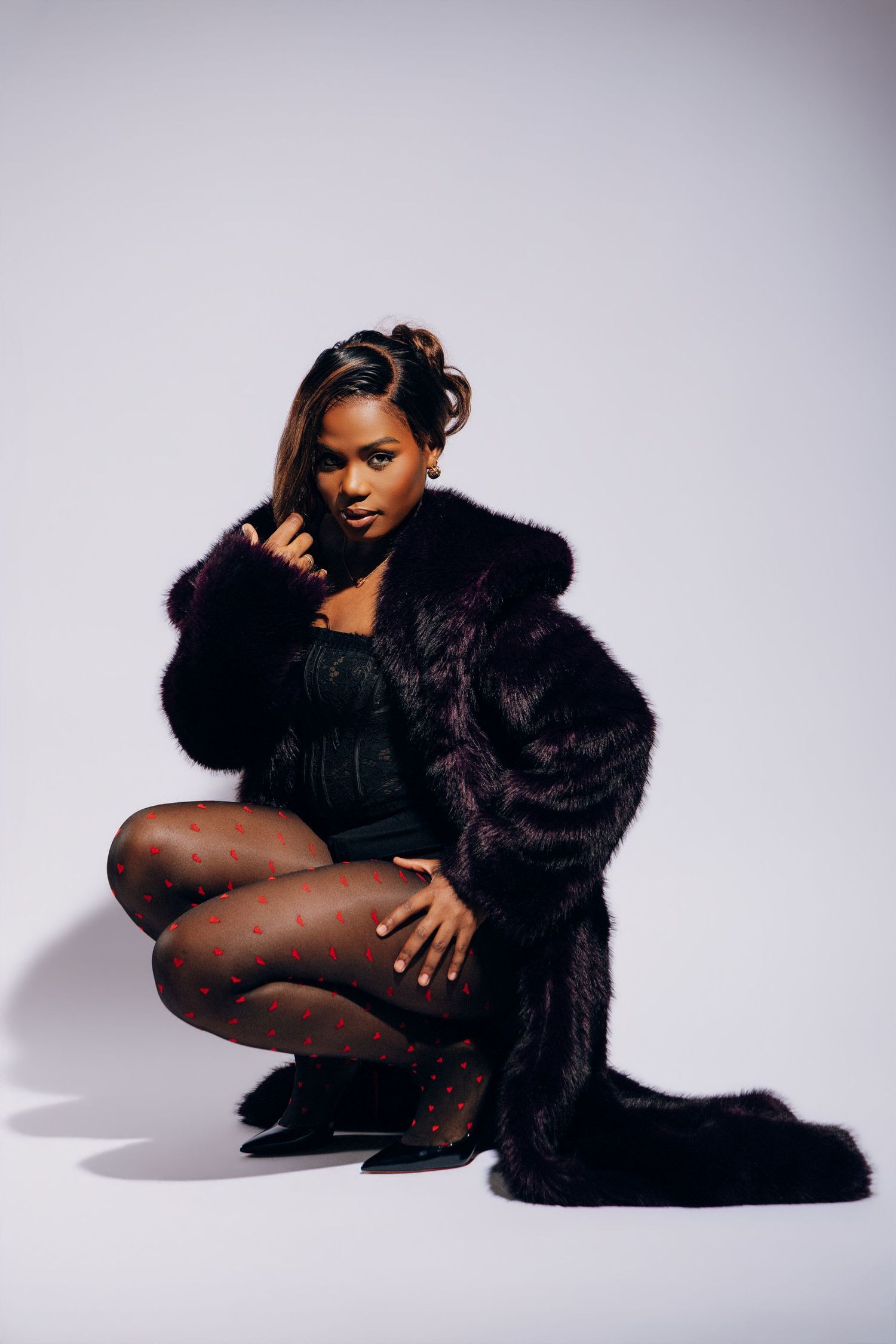 Woman in black fur coat and patterned tights posing confidently in a studio setting.