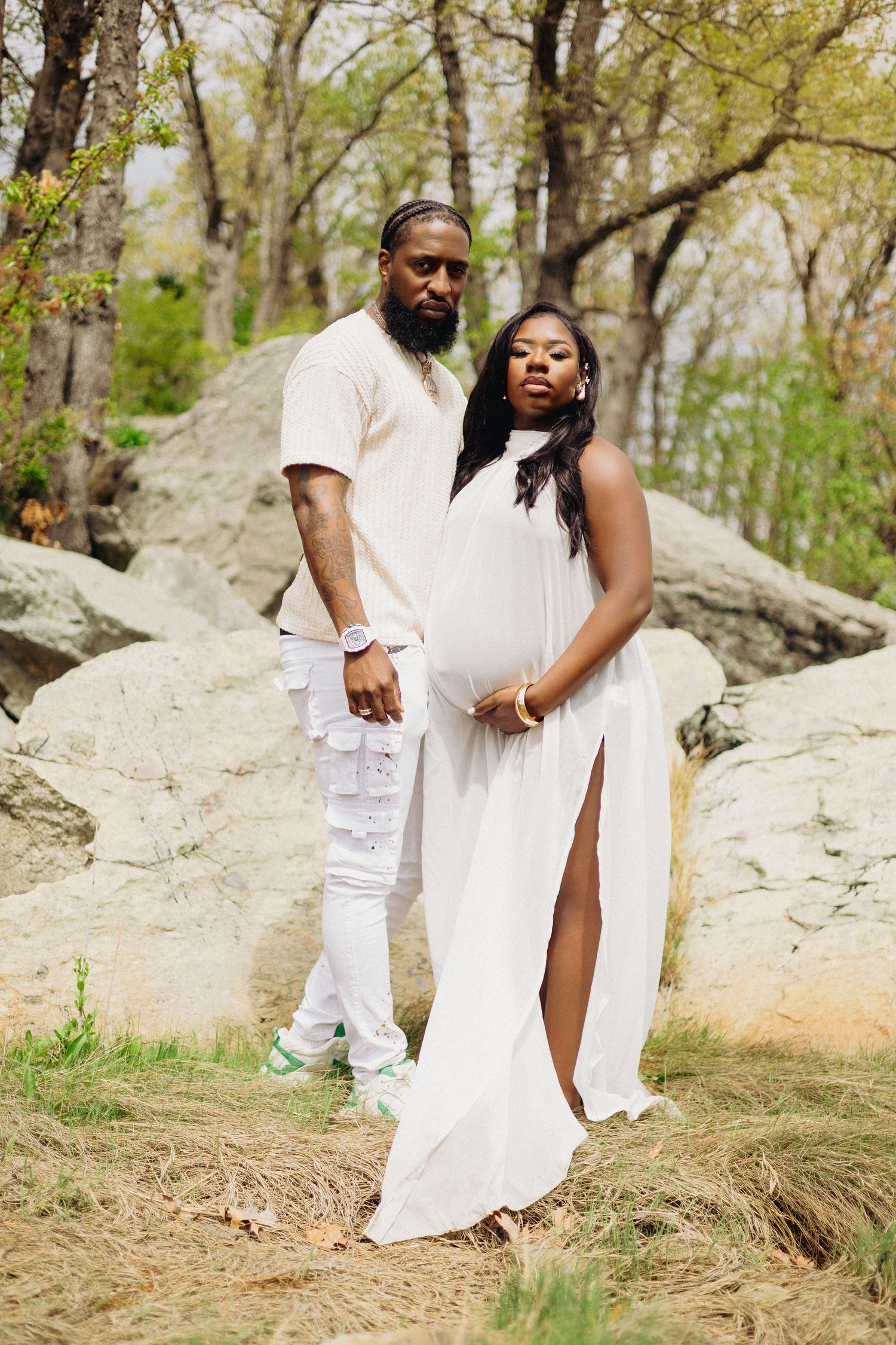 A couple in matching white outfits poses outdoors, the woman in a flowing maternity dress cradling her baby bump near rocks.