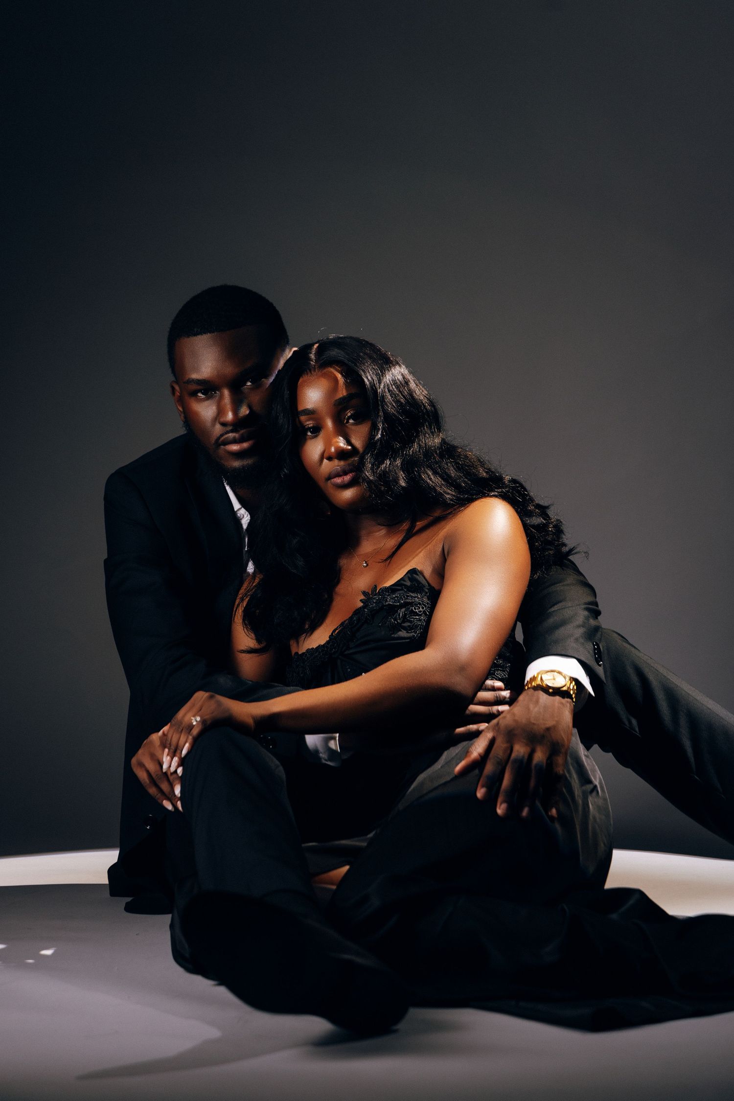 Elegant couple in black formal attire posing together in a studio, sitting closely in a sophisticated portrait.