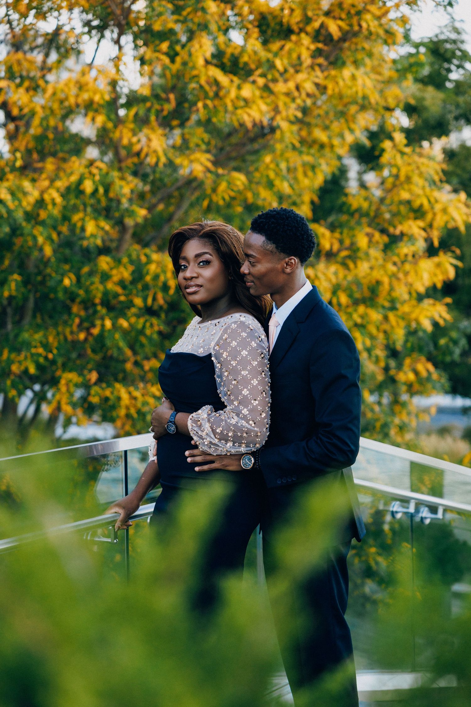 Elegant couple embracing on a balcony surrounded by autumn foliage, dressed in formal attire.