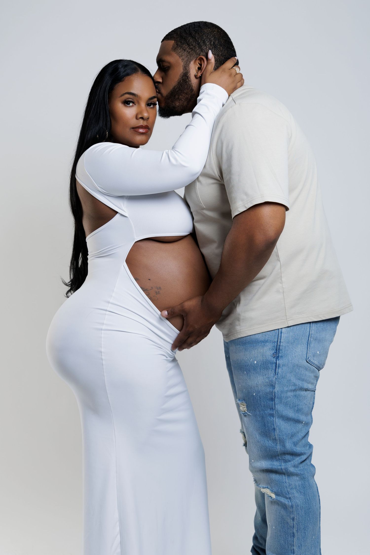 A couple poses for a maternity photo, the woman in a white cutout dress showing her baby bump, as her partner kisses her cheek.