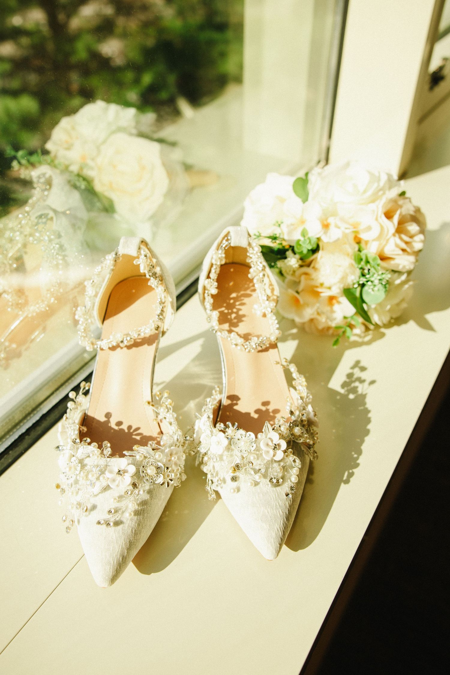 Elegant white bridal heels adorned with pearls and lace flowers displayed on a windowsill beside a soft floral bouquet.