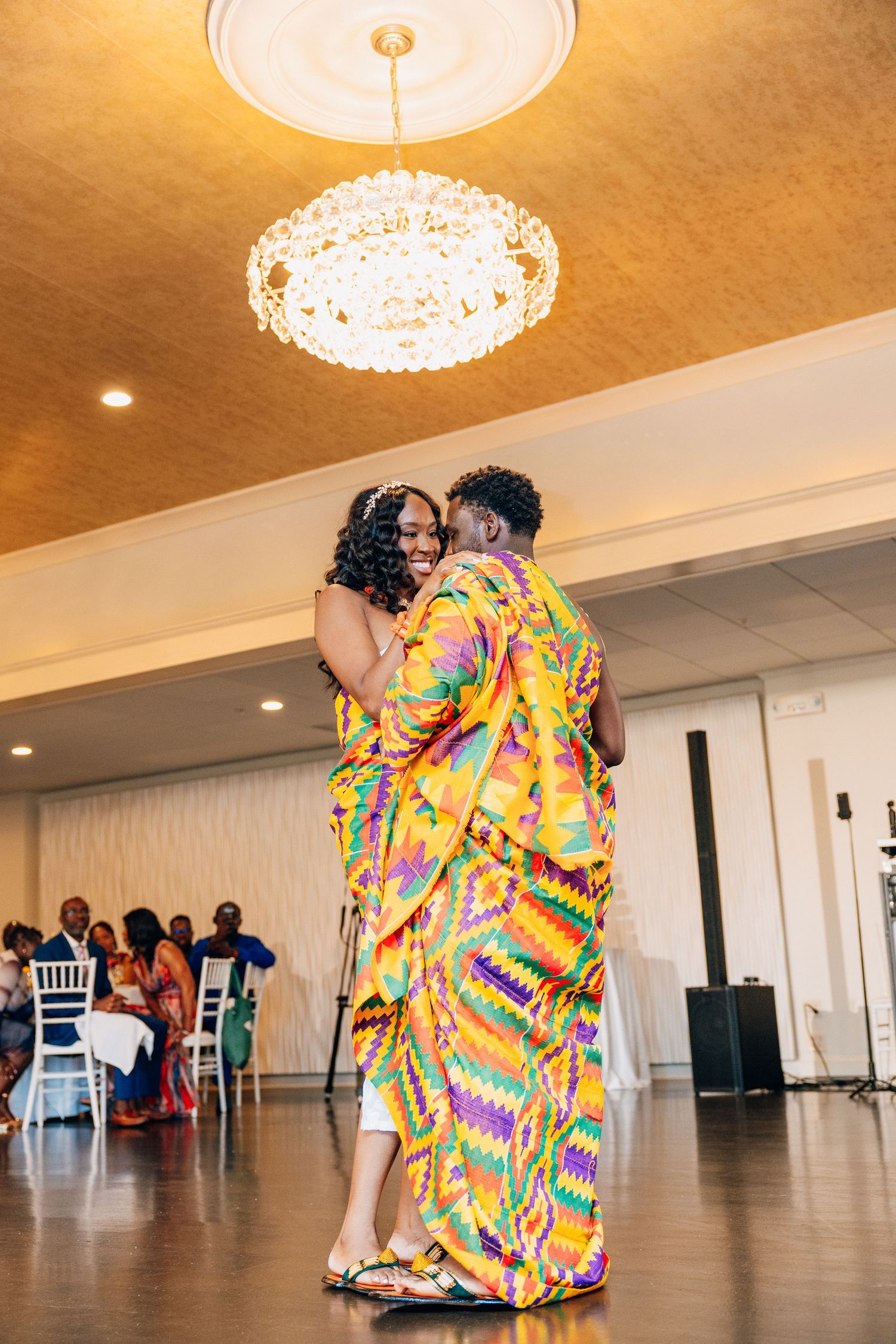 Bride and groom dancing joyfully in traditional Kente attire under chandelier.
