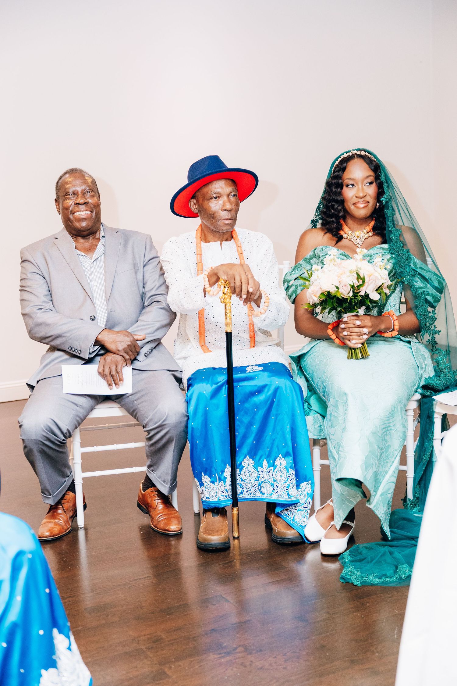 Bride in teal gown with elder in blue traditional attire and man in gray suit seated at African wedding ceremony.