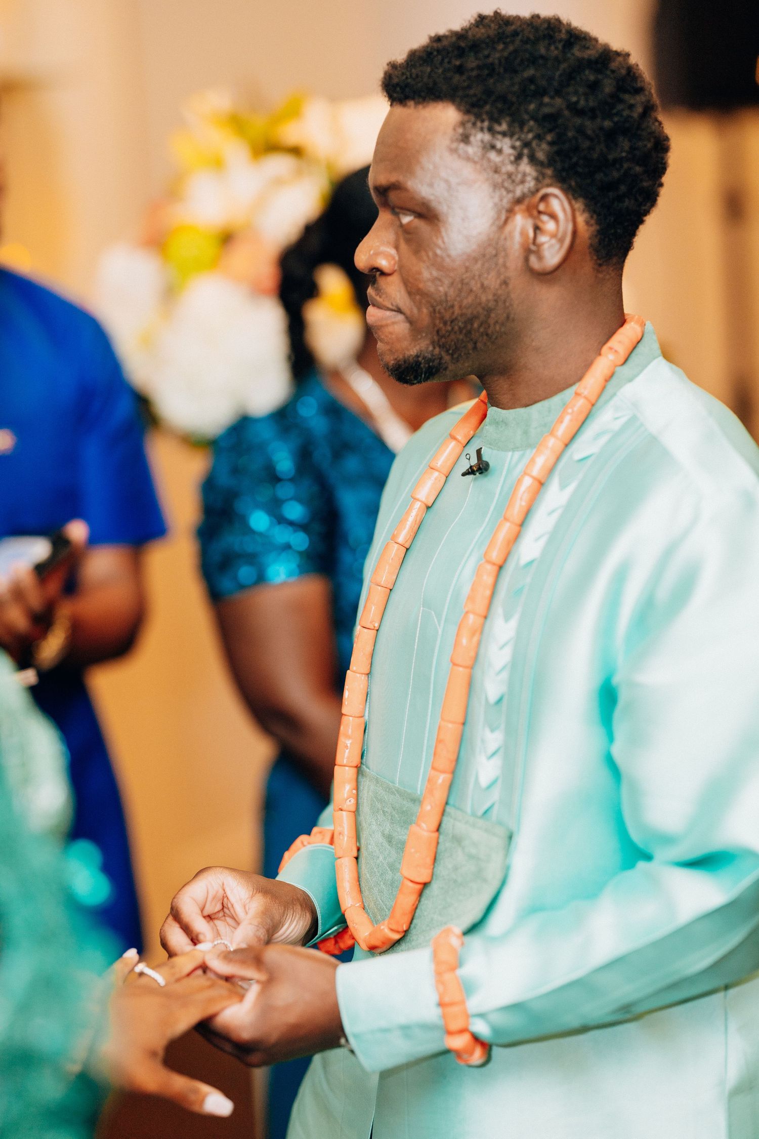 Groom in mint green traditional African attire with orange coral beaded necklace at wedding ceremony.