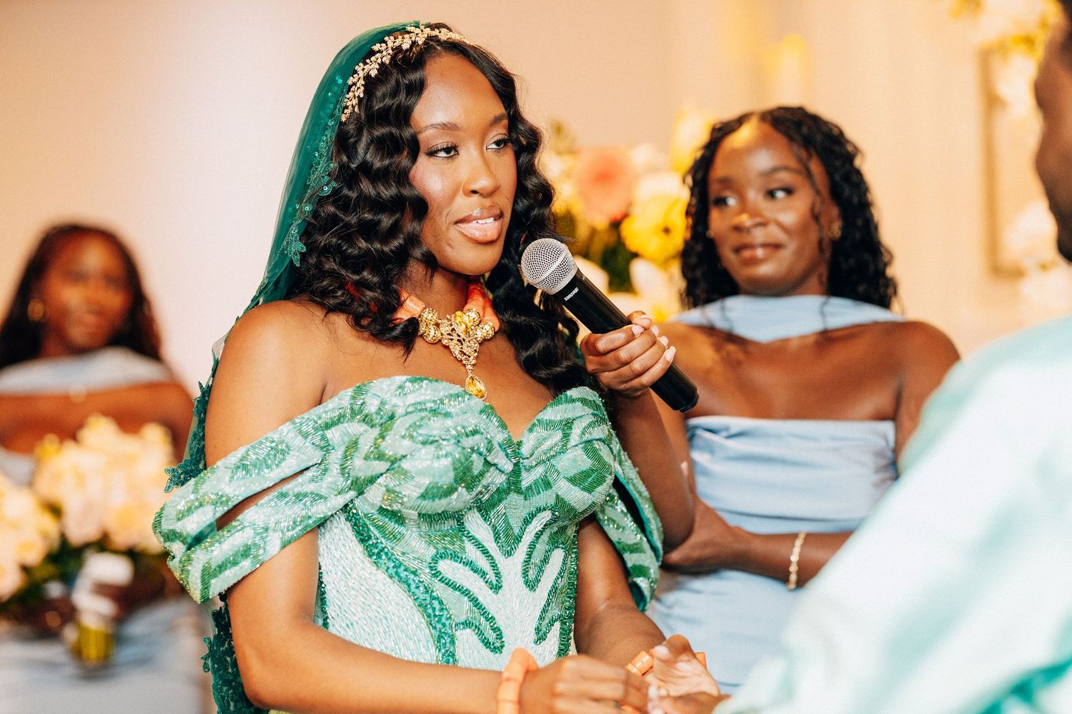Radiant bride in teal off-shoulder gown with gold headpiece speaks into microphone at wedding.