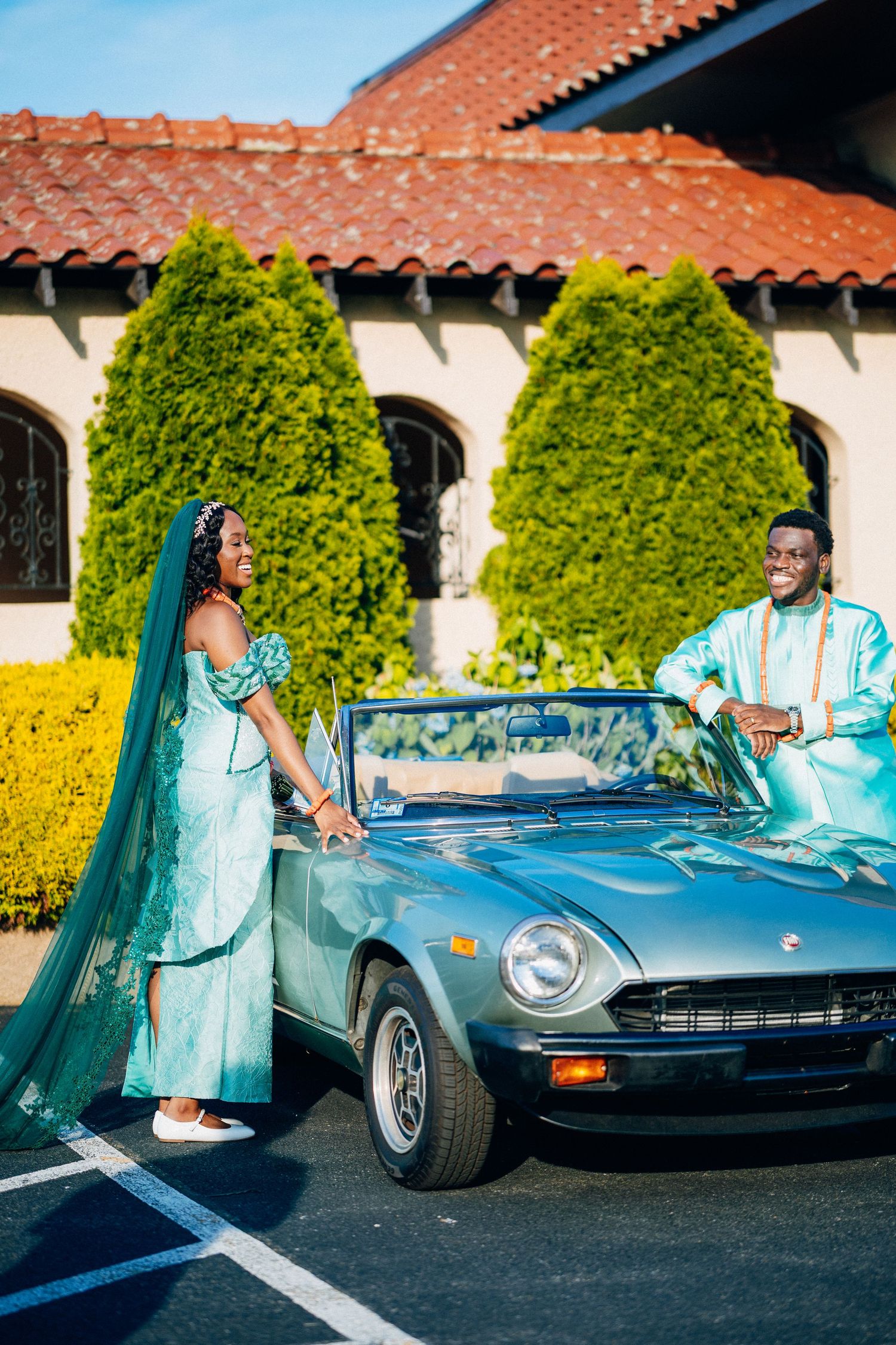 Nigerian wedding couple in matching teal traditional attire posing with vintage silver convertible.