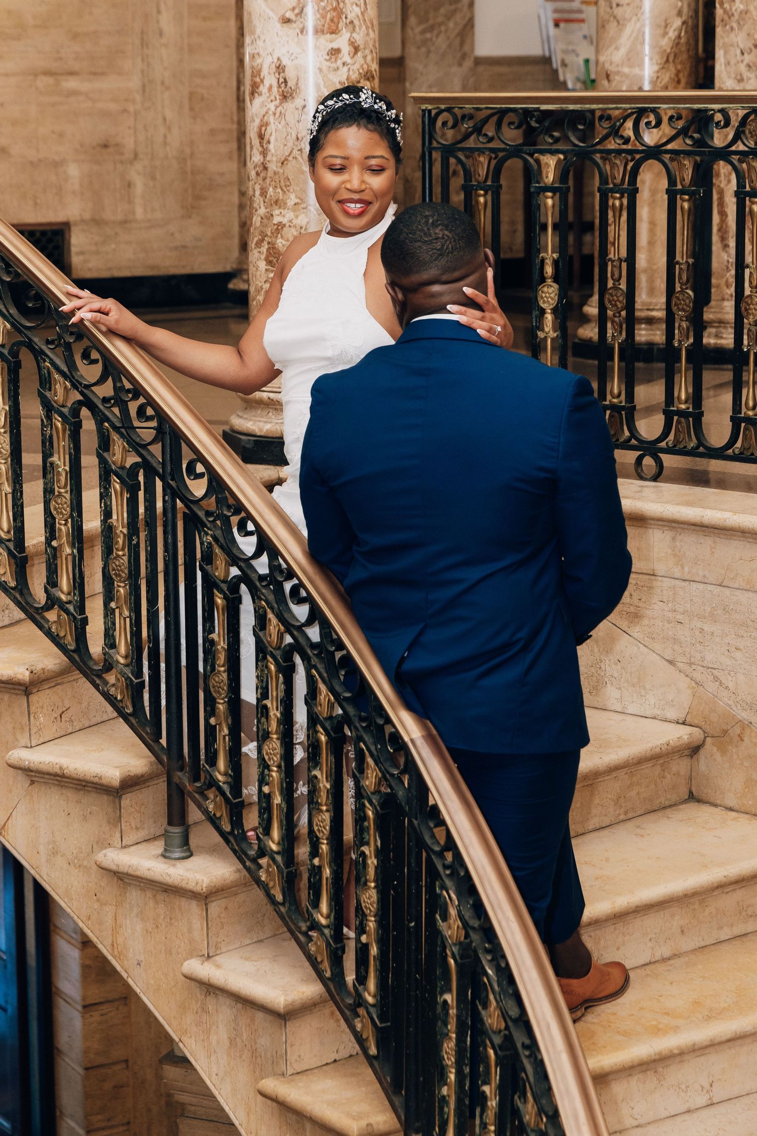 Wedding couple on ornate curved staircase, bride in white backless gown smiling at groom in navy suit.