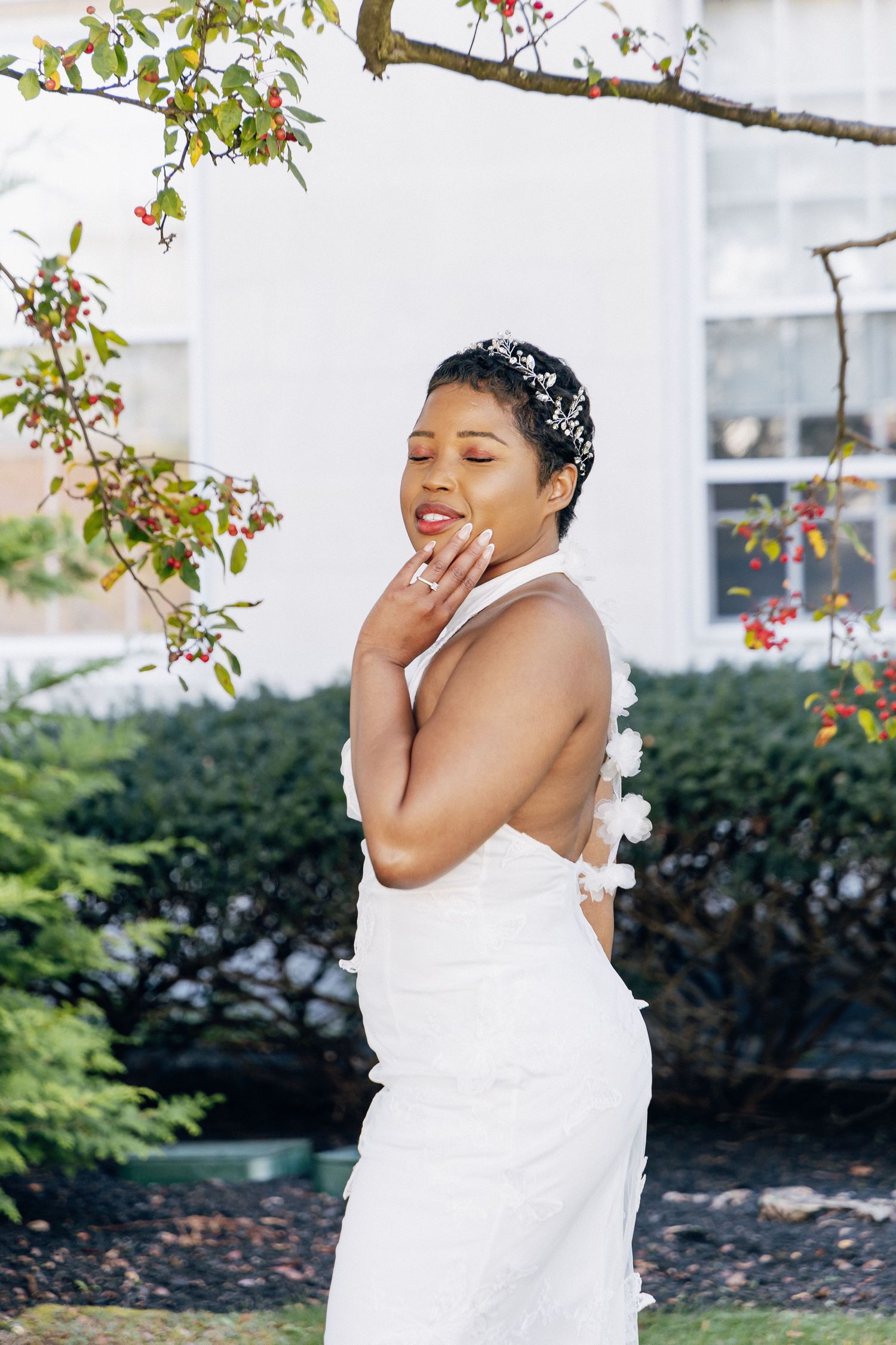 Bride in backless white halter wedding gown with floral details posing outdoors near berry tree.