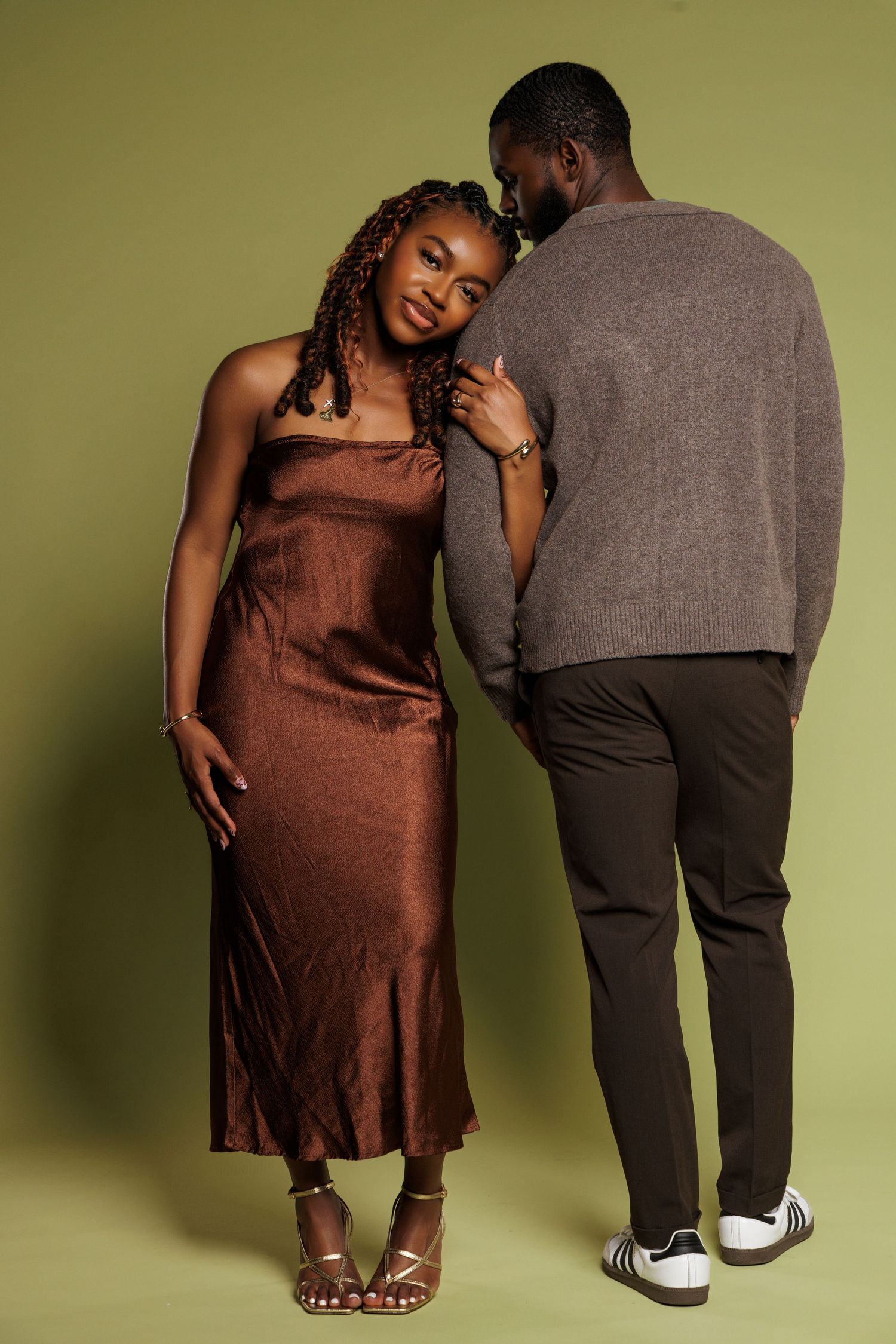 Full length shot of a Black couple in elegant brown satin dress and gray cardigan posing on a green background.
