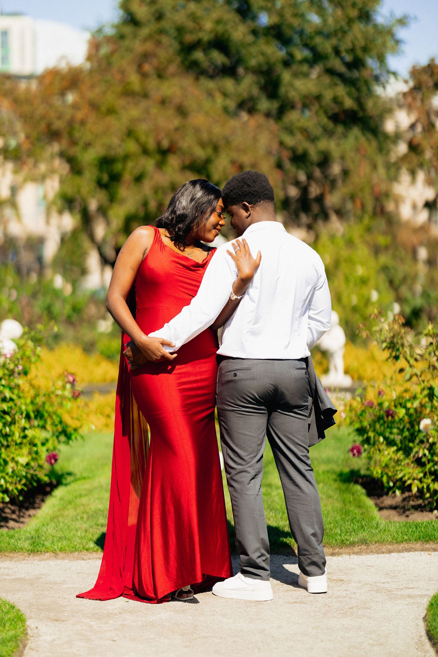 Romantic couple nearly kissing in colorful garden, woman in red gown and man in gray pants.