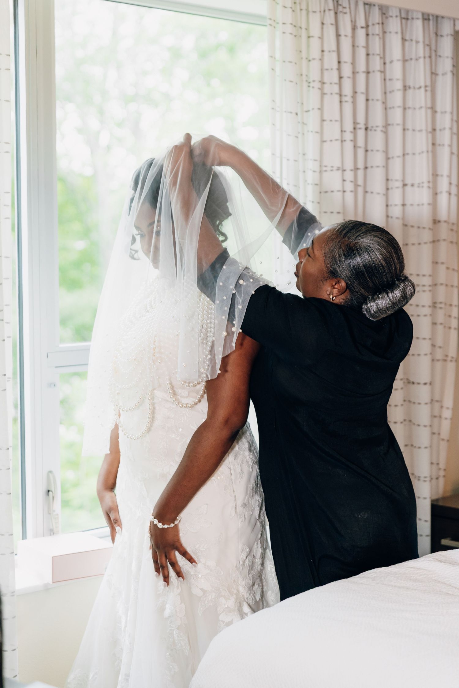 Mother helping Black bride adjust pearl-trimmed wedding veil over lace gown by window.