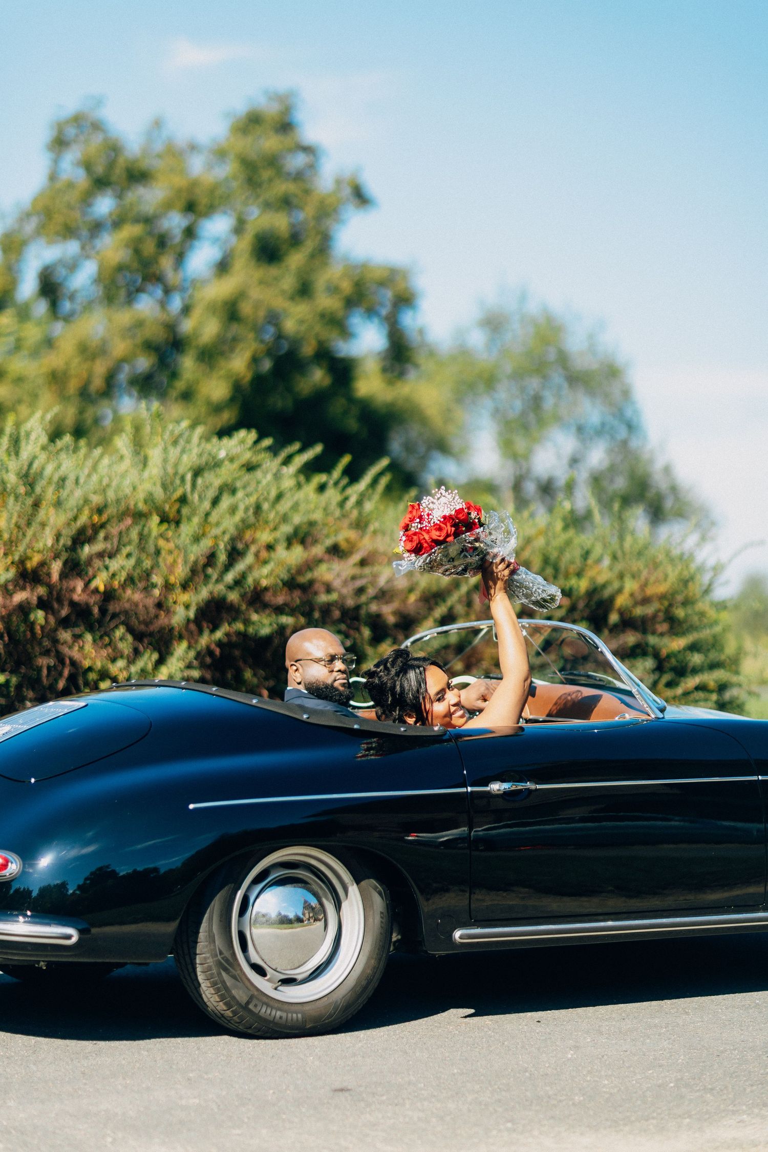 Bride raises red bouquet in classic black Porsche 356 convertible with groom driving on sunny day.