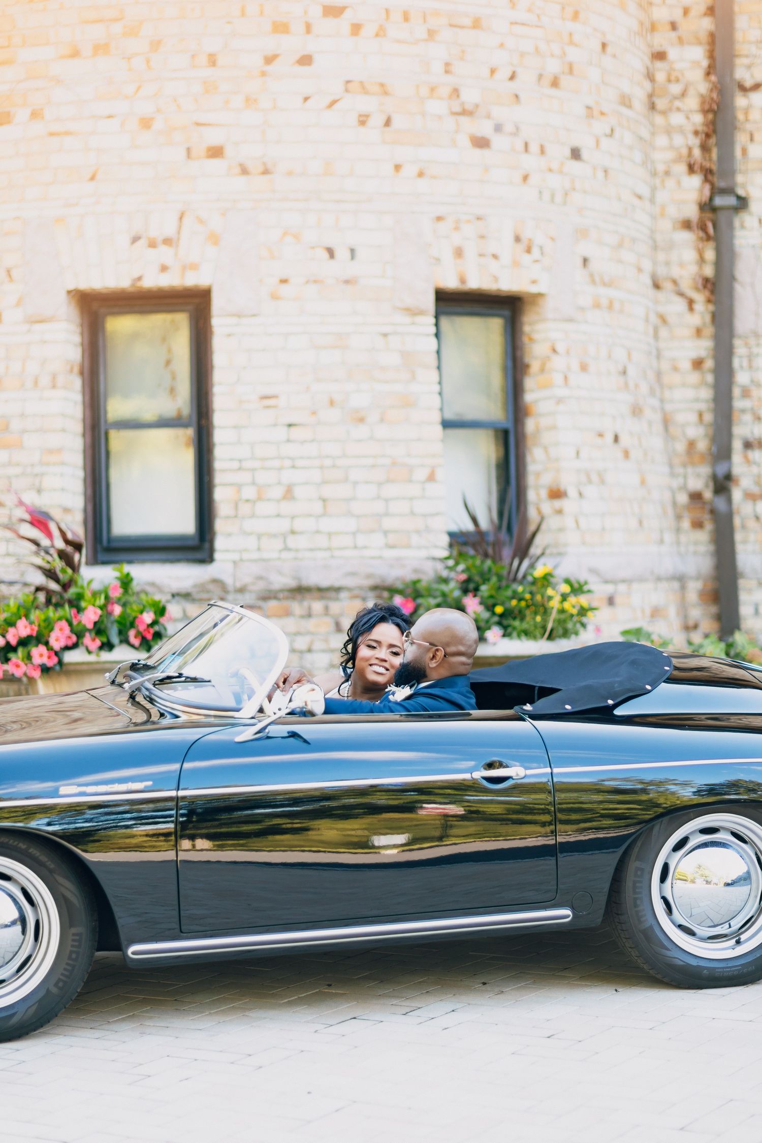 Newlywed couple sitting together in a classic black convertible sports car, smiling at each other on their wedding day.