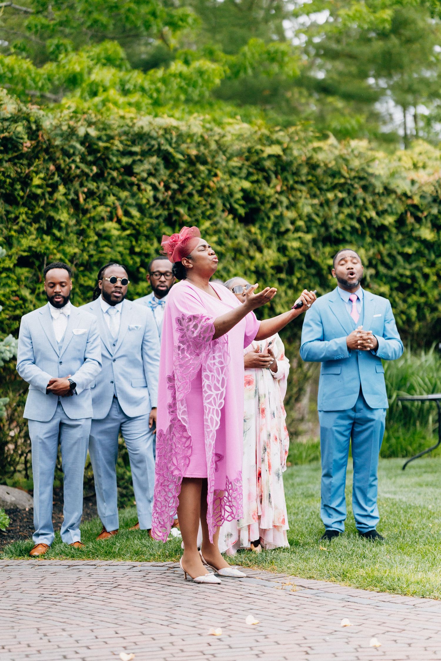 Woman in pink lace dress with arms raised singing at outdoor wedding with groomsmen behind her.