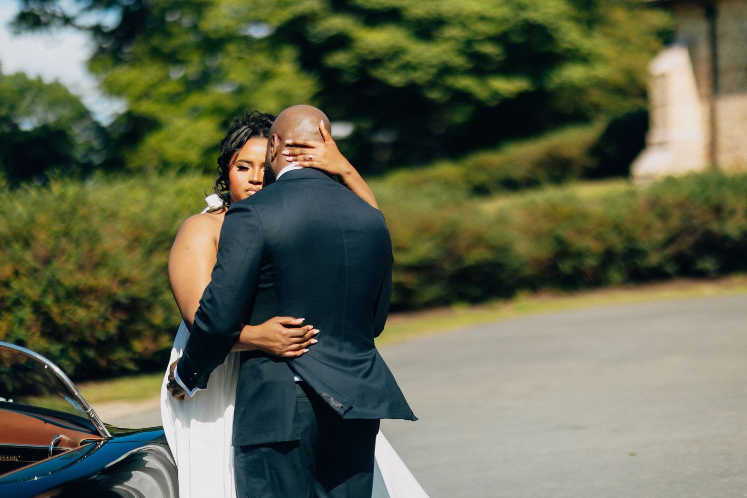 African American bride and groom sharing a tender embrace beside a vintage blue convertible car.