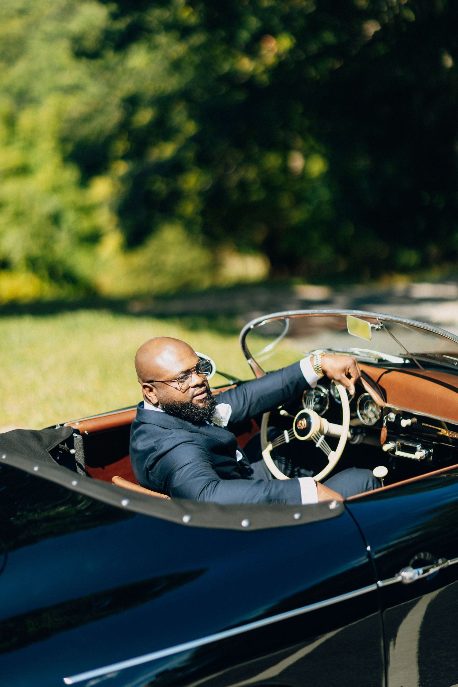 Stylish groom in navy suit drives vintage black convertible car outdoors on sunny day.