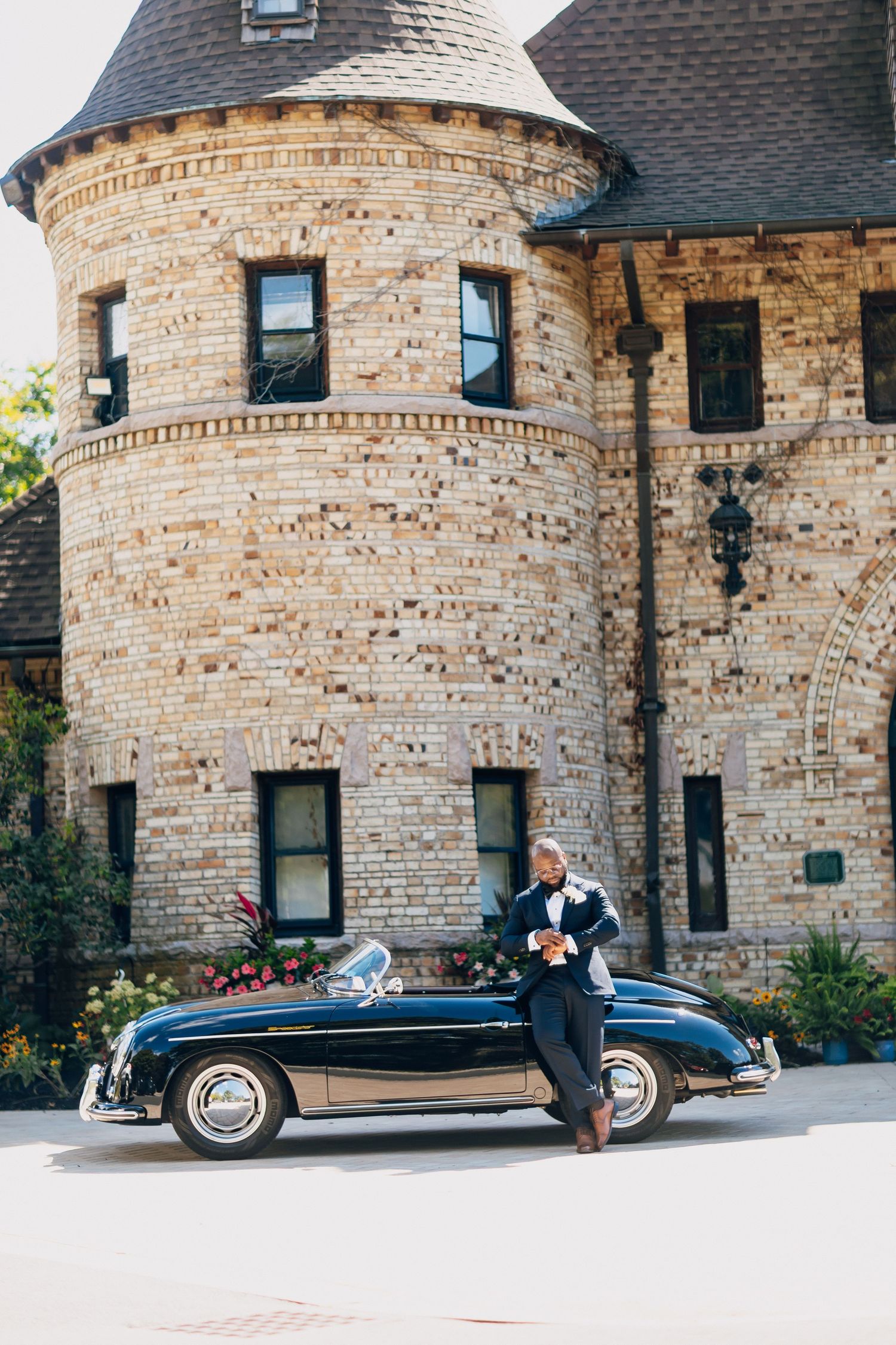 Dapper groom in navy tuxedo leans against vintage black Porsche before a stone castle building.