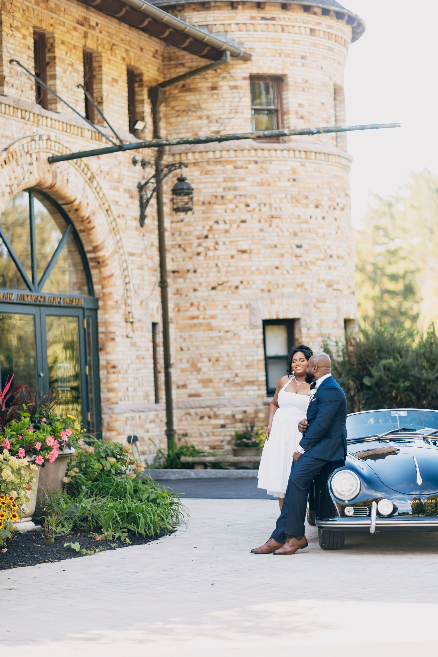 Wedding couple poses beside a classic black Porsche in front of an elegant brick tower venue.