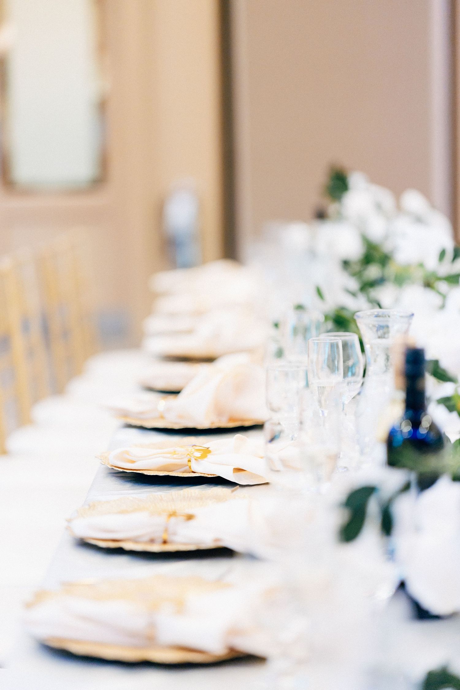 Elegant wedding reception table set with gold charger plates, white napkins, crystal glasses, and floral greenery centerpiece.