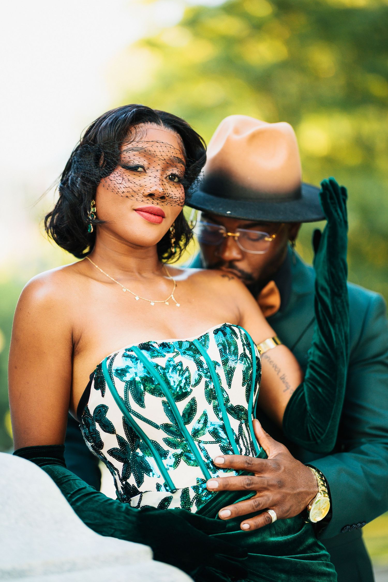 Elegant woman in green sequined corset gown with veil, embraced by man in teal suit.