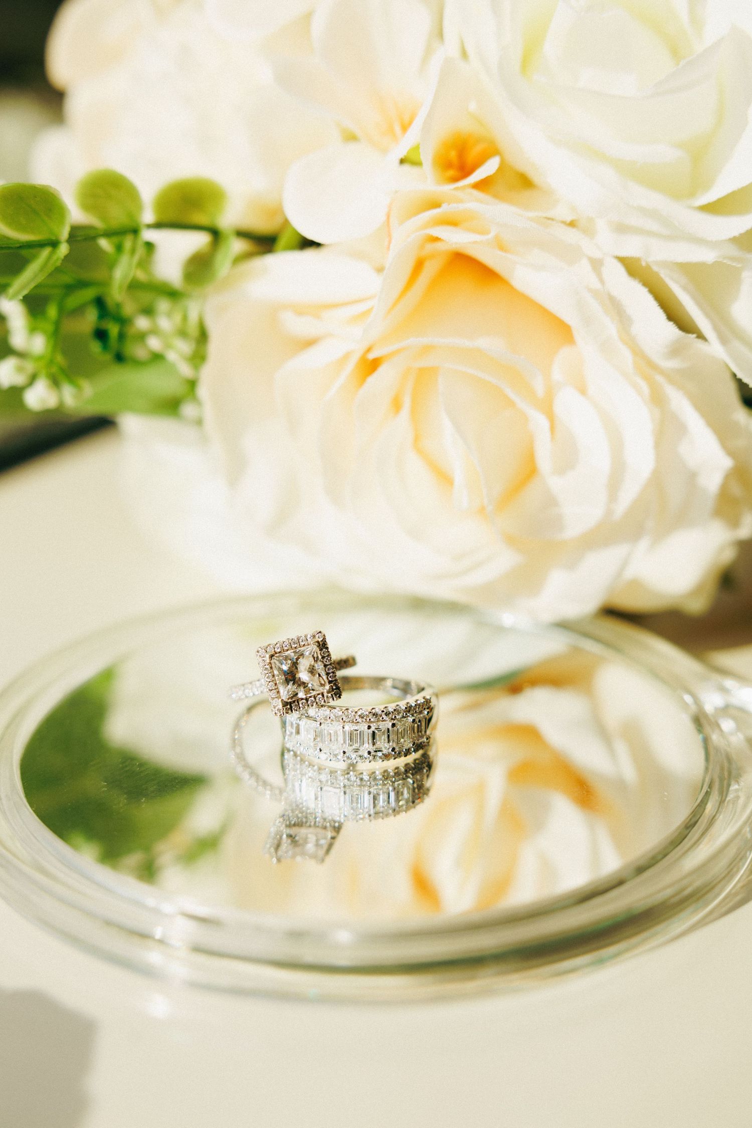 Diamond engagement ring and wedding band reflected on glass dish beside white roses bouquet.