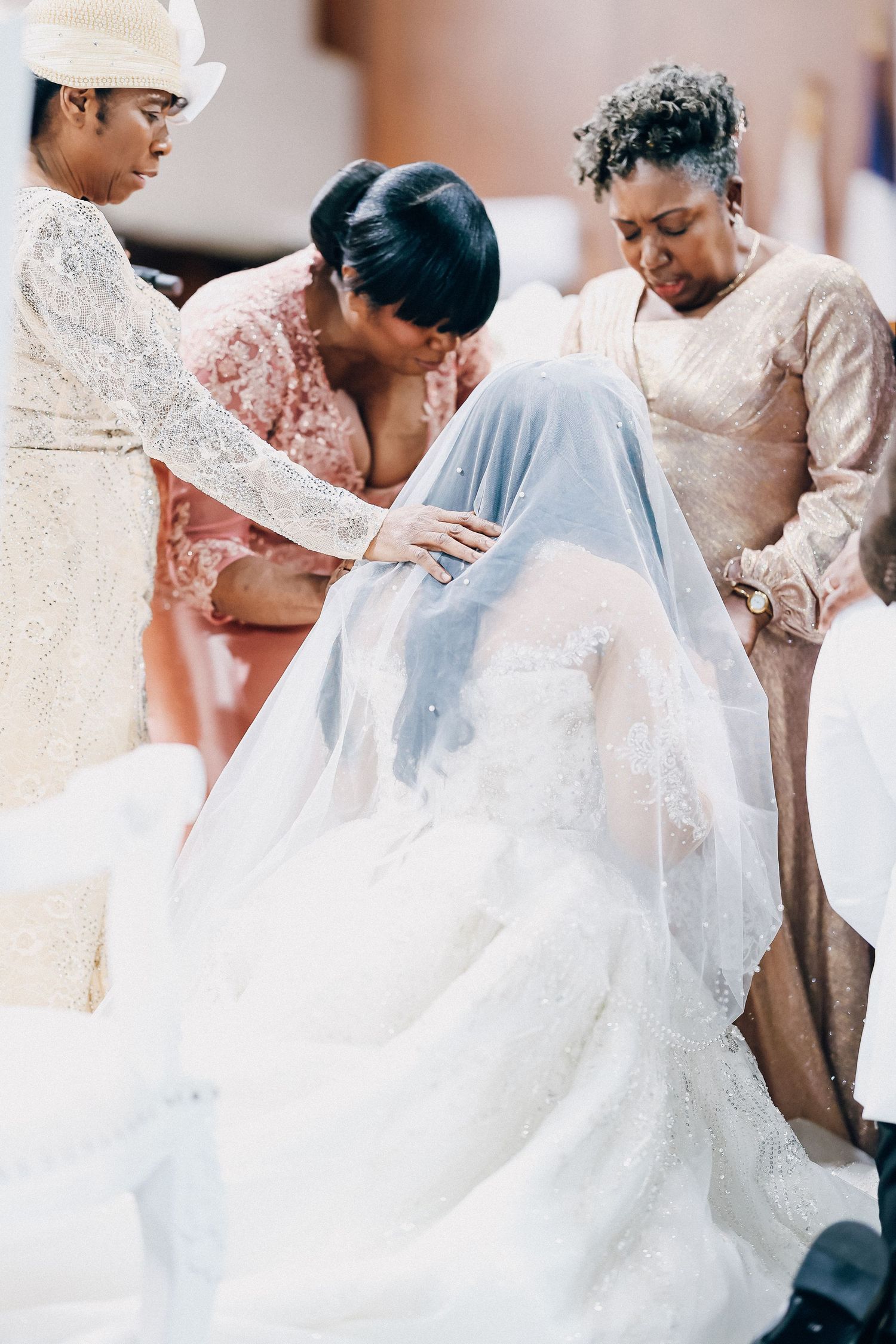 Bride kneeling in white gown receiving blessing from three women during emotional wedding ceremony prayer.