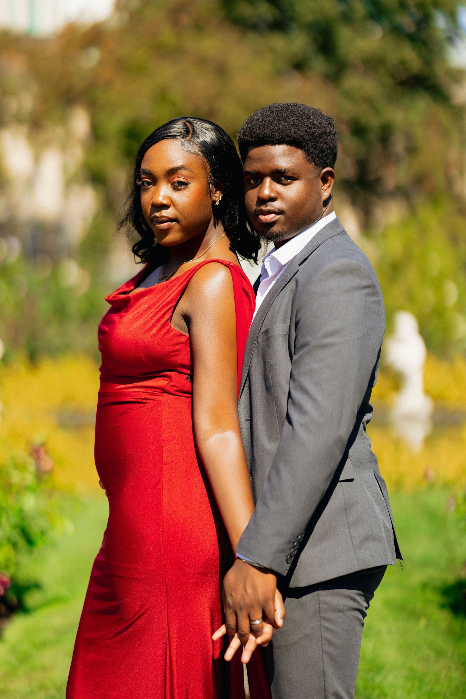 A young Black couple poses in a garden, woman in red dress and man in gray suit, holding hands.