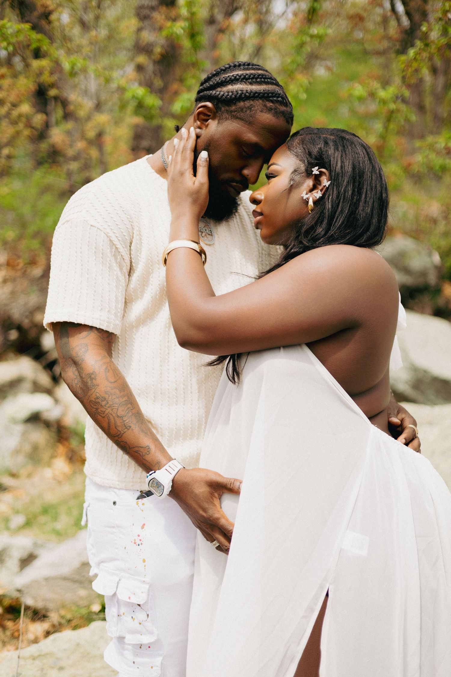 A couple in white outfits share a tender embrace outdoors, with the woman resting her hand on the man's face.