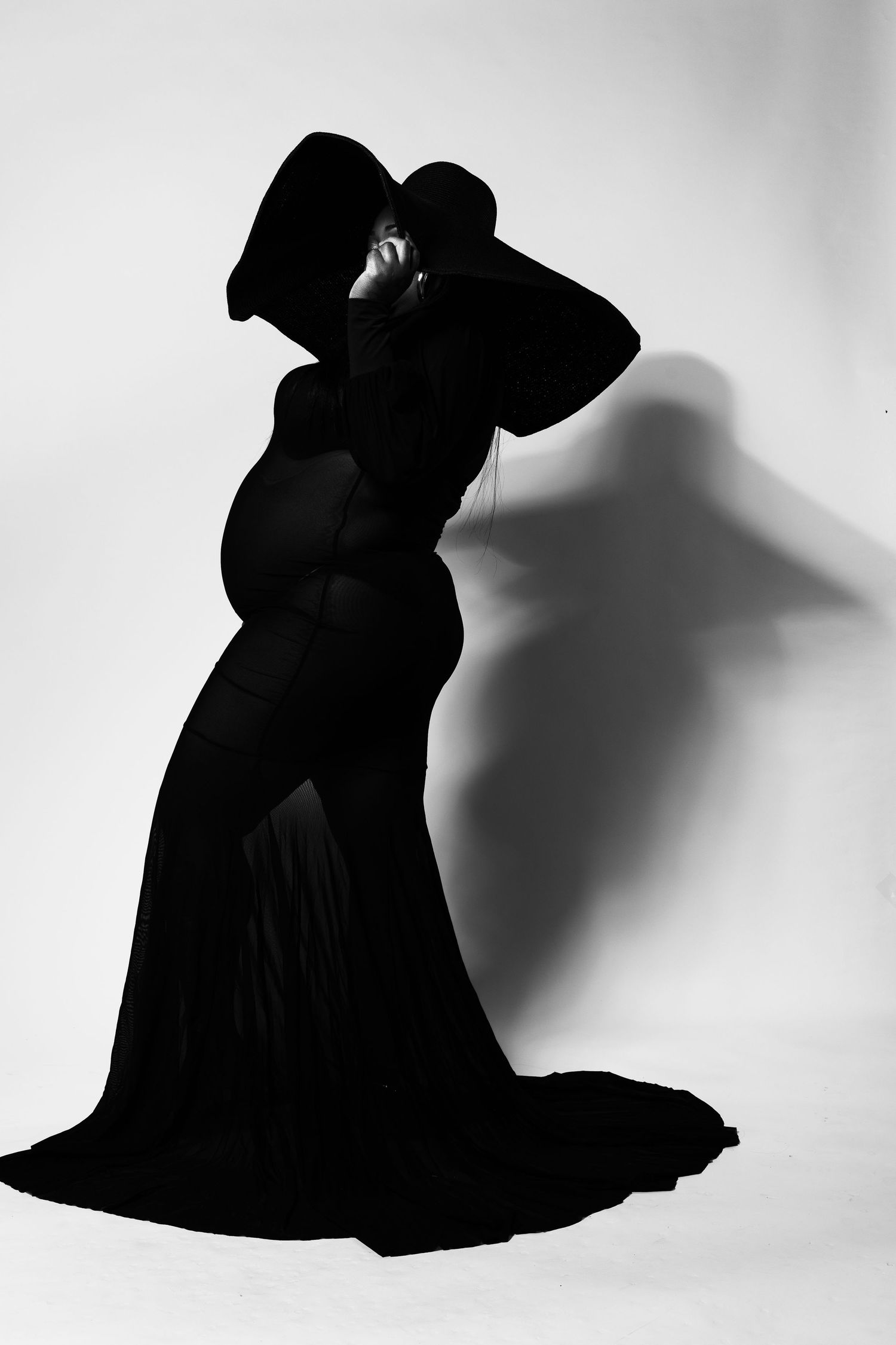Dramatic black and white silhouette of a pregnant woman in elegant black gown and wide-brim hat.