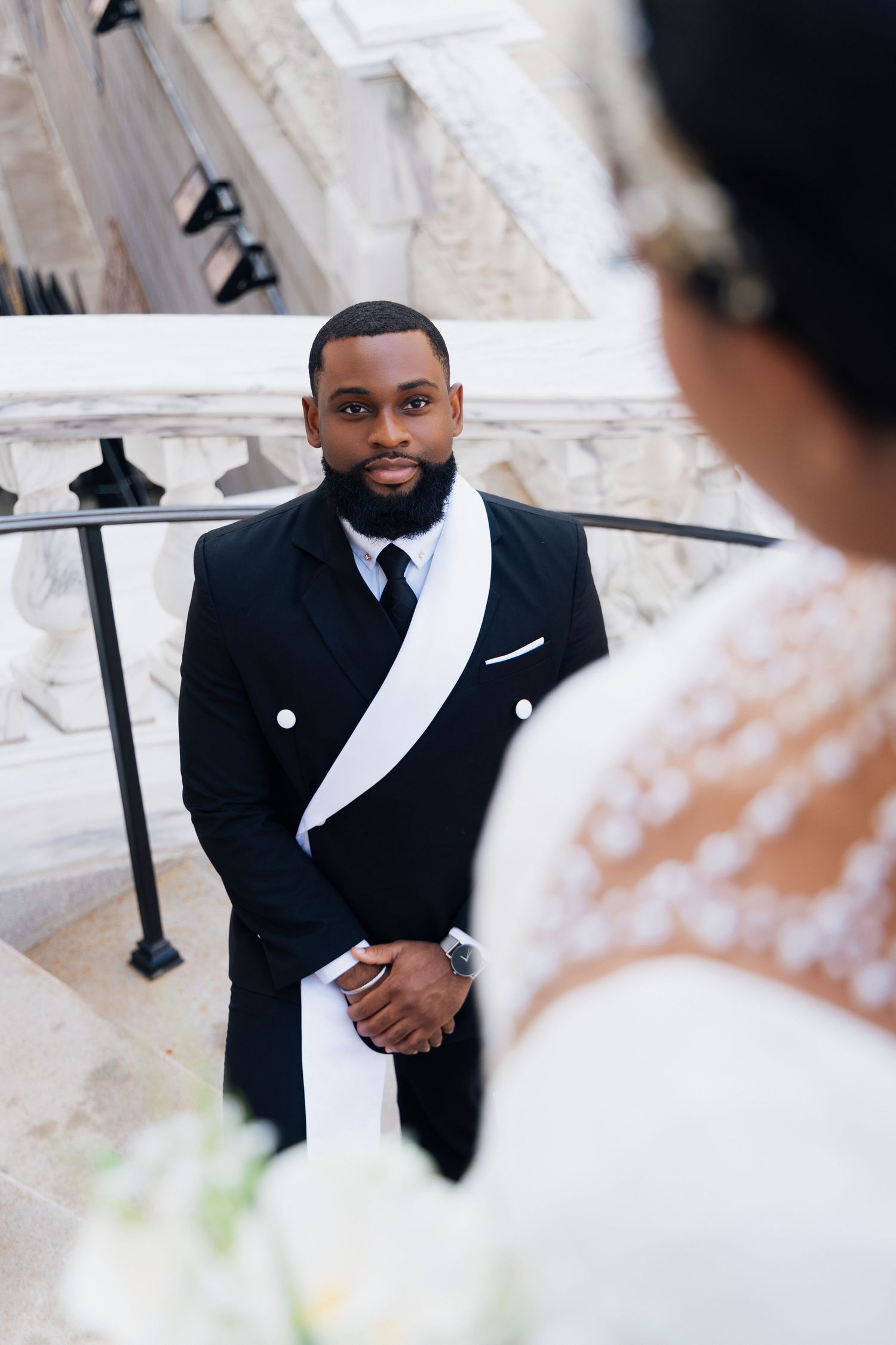 Groom in black tuxedo with white sash gazes at bride on marble staircase during elegant wedding ceremony.