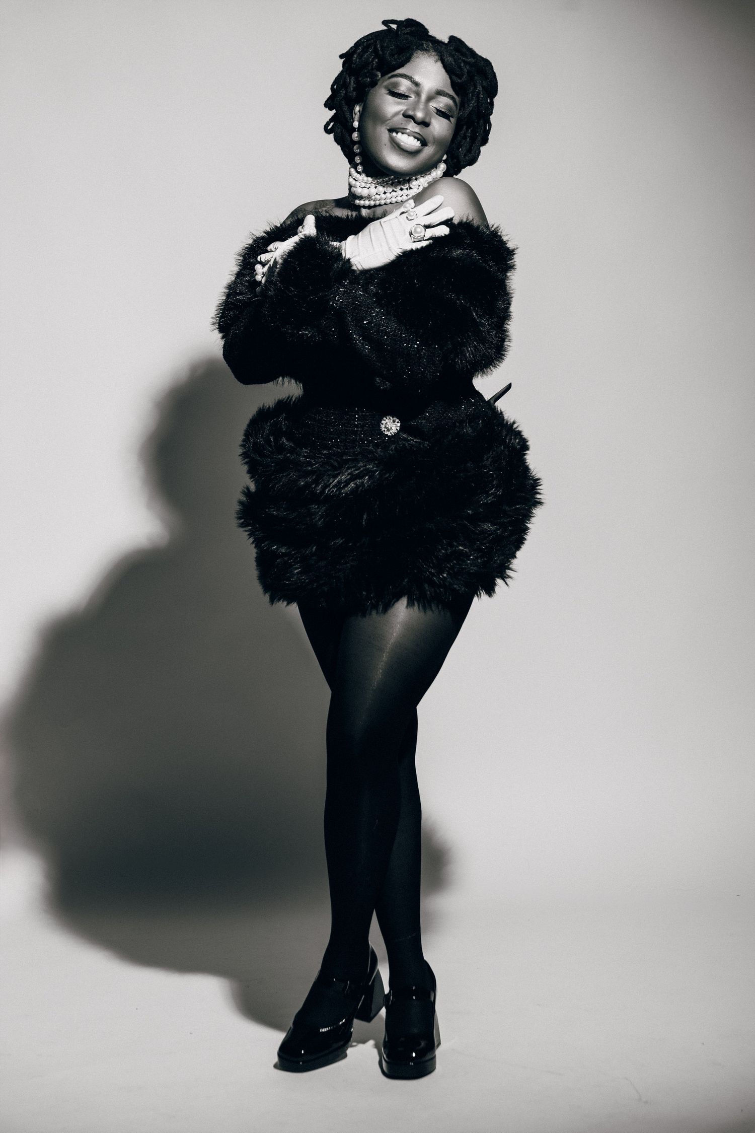 Black and white photo of a smiling woman in a black fur coat, pearl necklace, and heels posing confidently.