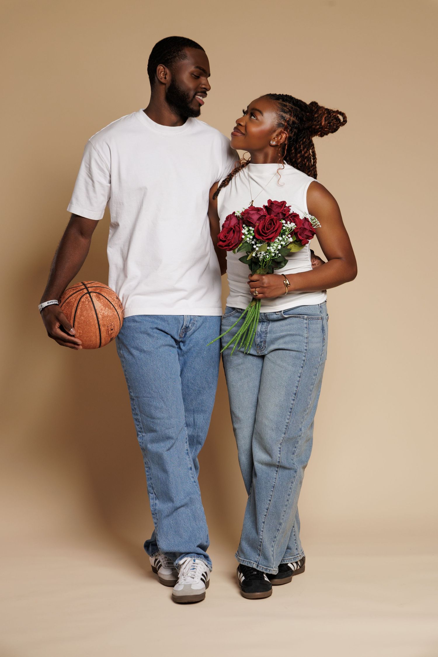 A couple in white tees and jeans, man holding basketball, woman holding red roses bouquet, posing against beige background.
